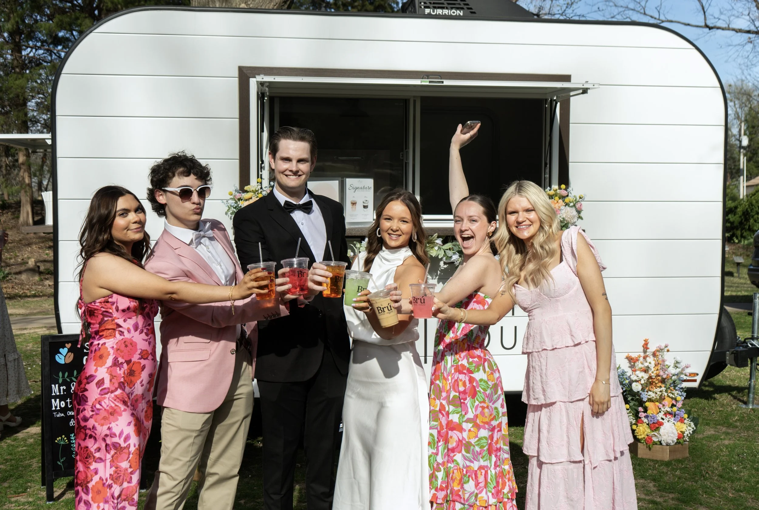 Group of friends celebrating outdoors in front of a mobile coffee stand, holding up drinks and smiling. They are dressed in colorful, semi-formal attire.