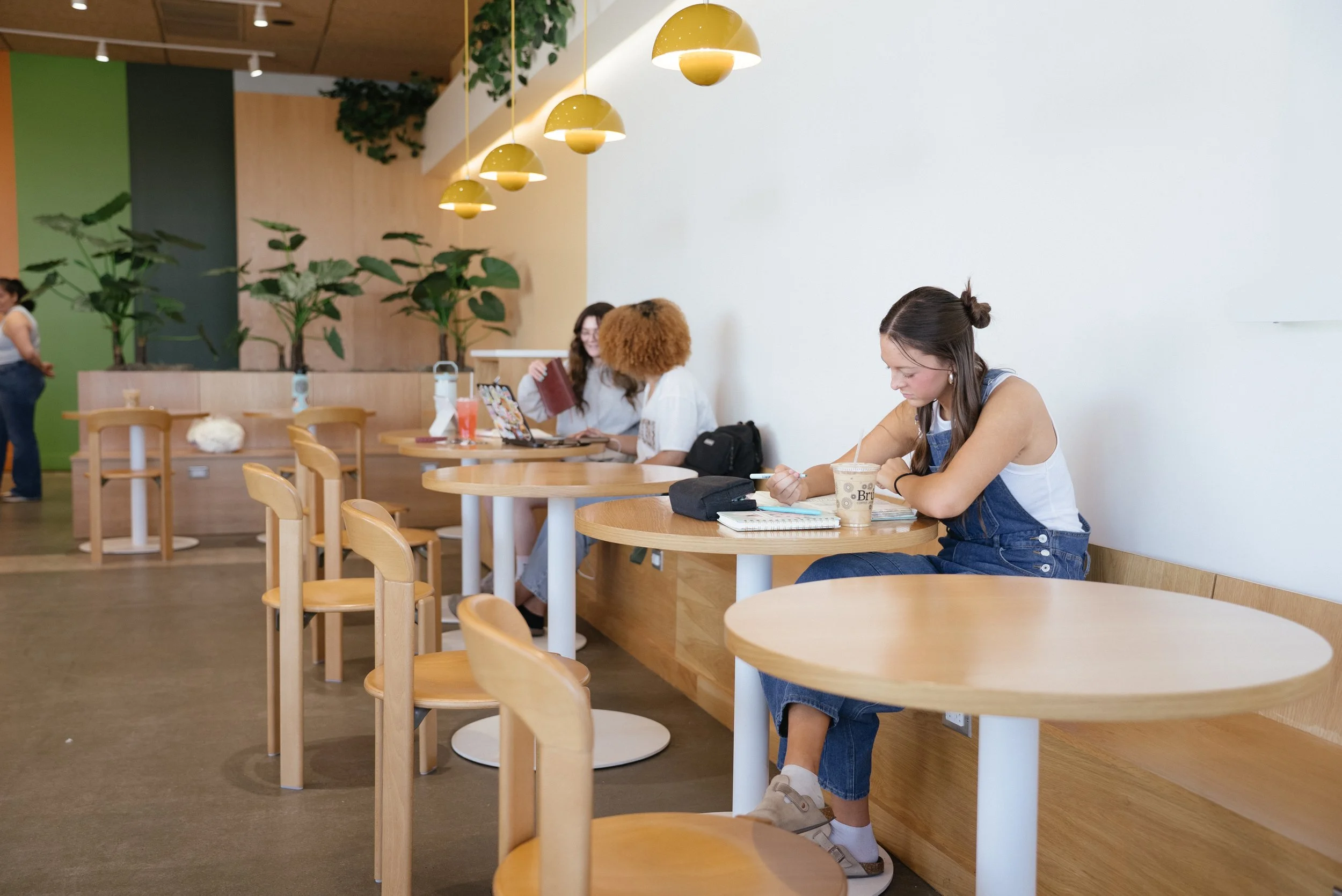 Young woman sitting at a wooden table in a modern cafe, writing in a notebook with a drink beside her. Several other women are seated at tables, some reading or using laptops, and a woman is standing near a green wall in the background. The cafe has hanging yellow pendant lights and potted plants.