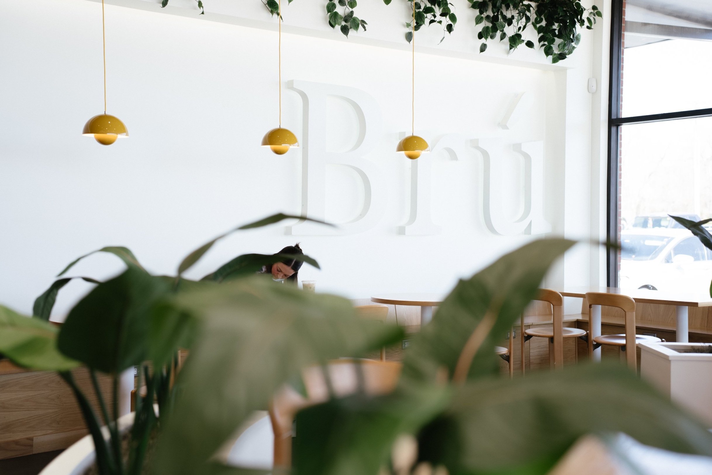 Interior of a modern cafe with a large white wall featuring the word 'Bru', hanging yellow pendant lights, wooden chairs and tables, a large window, and a partially obscured woman enjoying a beverage.
