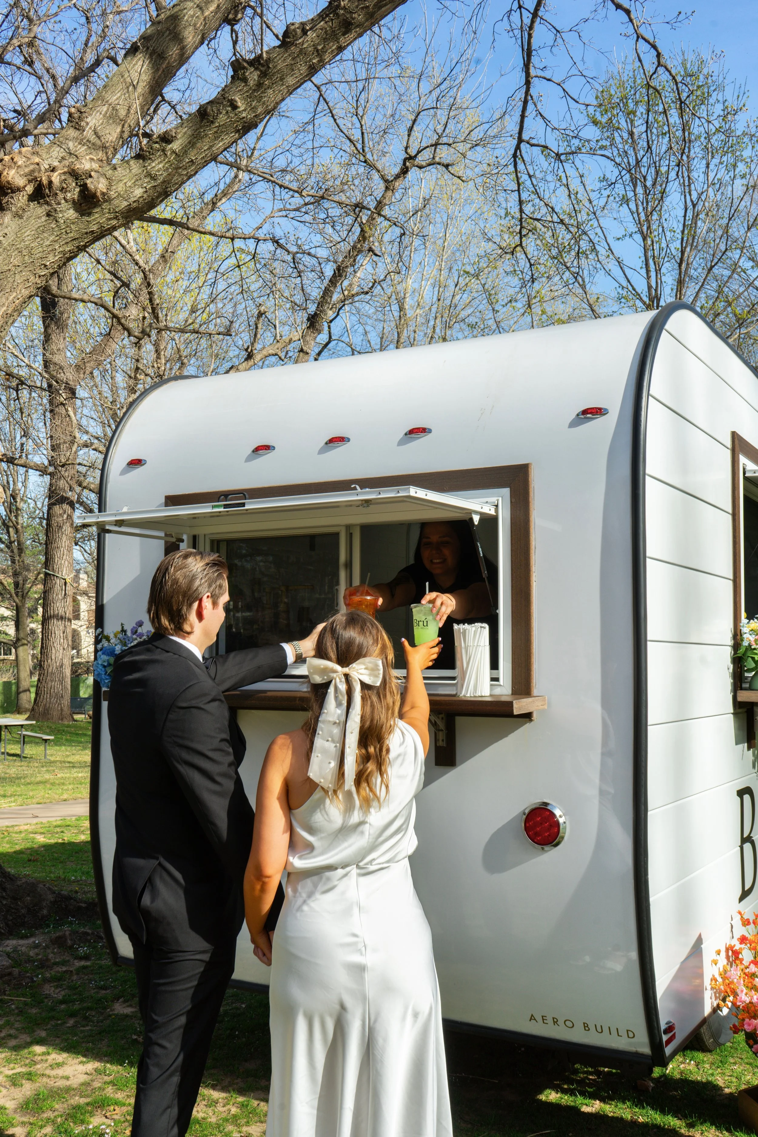 People at a food truck counter outdoors on a sunny day, with trees and a blue sky in the background.