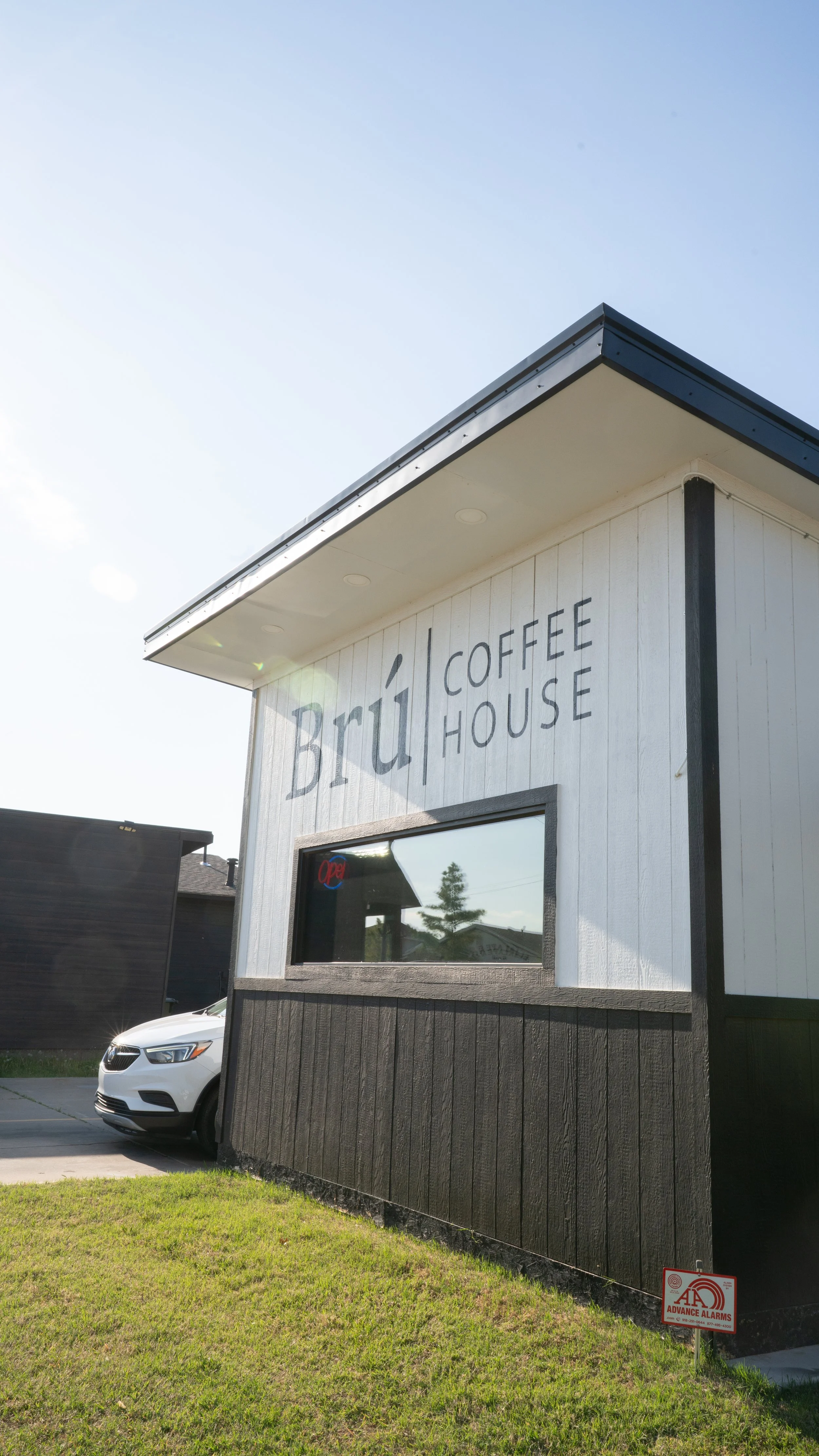 Exterior of a small coffee shop with a sign that reads 'Bru Coffee House'; white building with black accents, window reflecting trees and sky, car parked nearby, and a small sign in grass indicating alarm system.