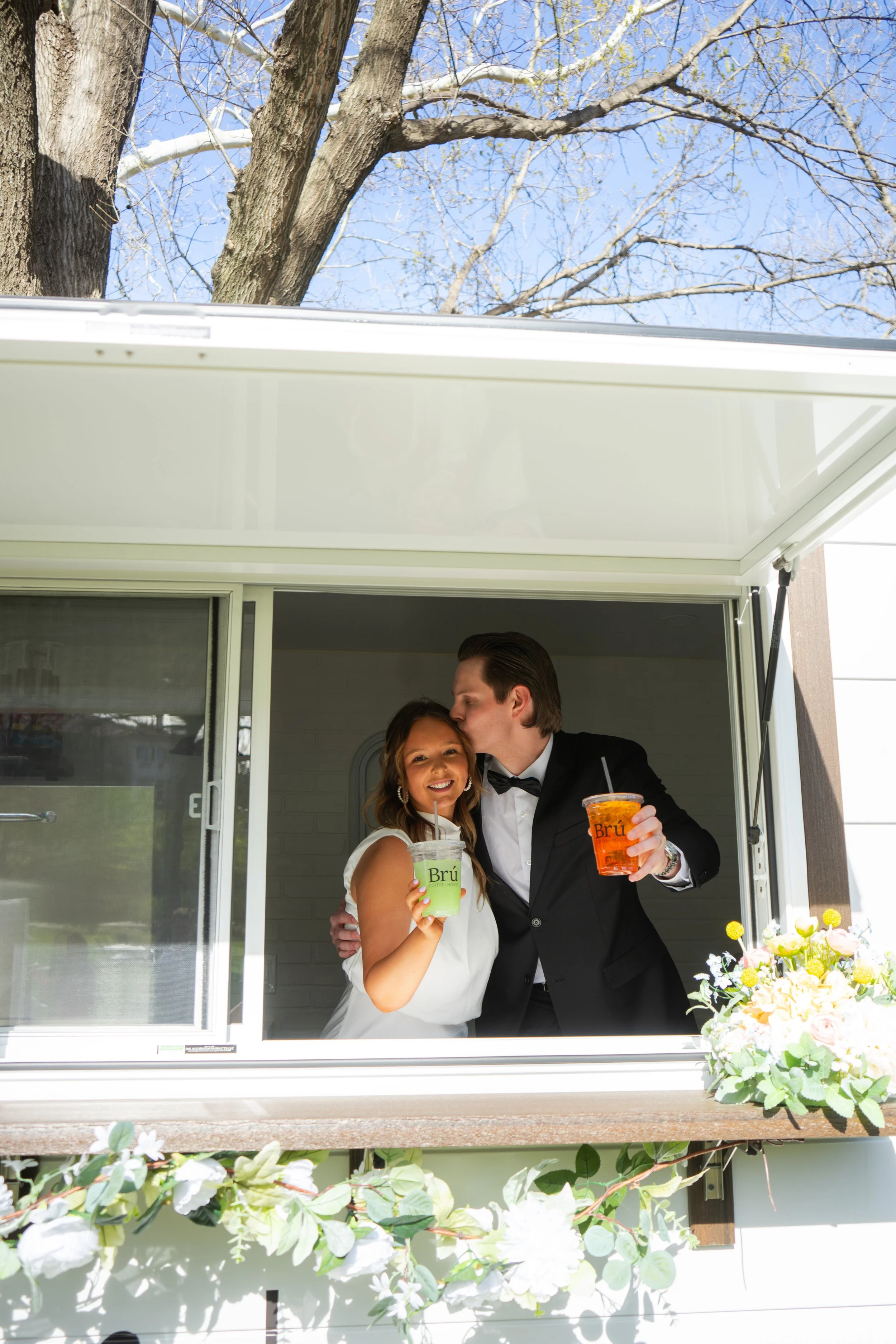 A man and woman in formal attire sharing a tender moment at a window, holding colorful drinks, surrounded by flowers.