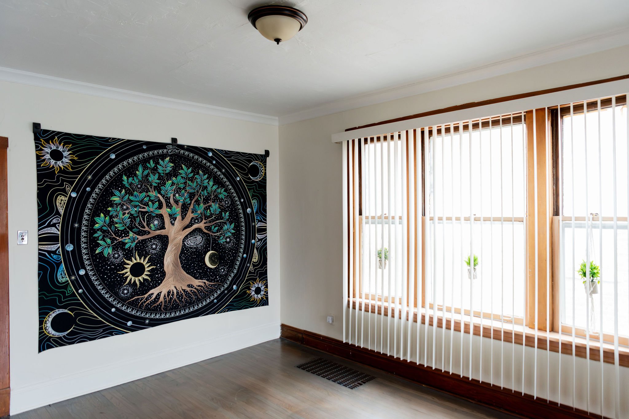 Living room with a large black and white tapestry featuring a tree and celestial designs hanging on the wall, next to a window with vertical blinds.