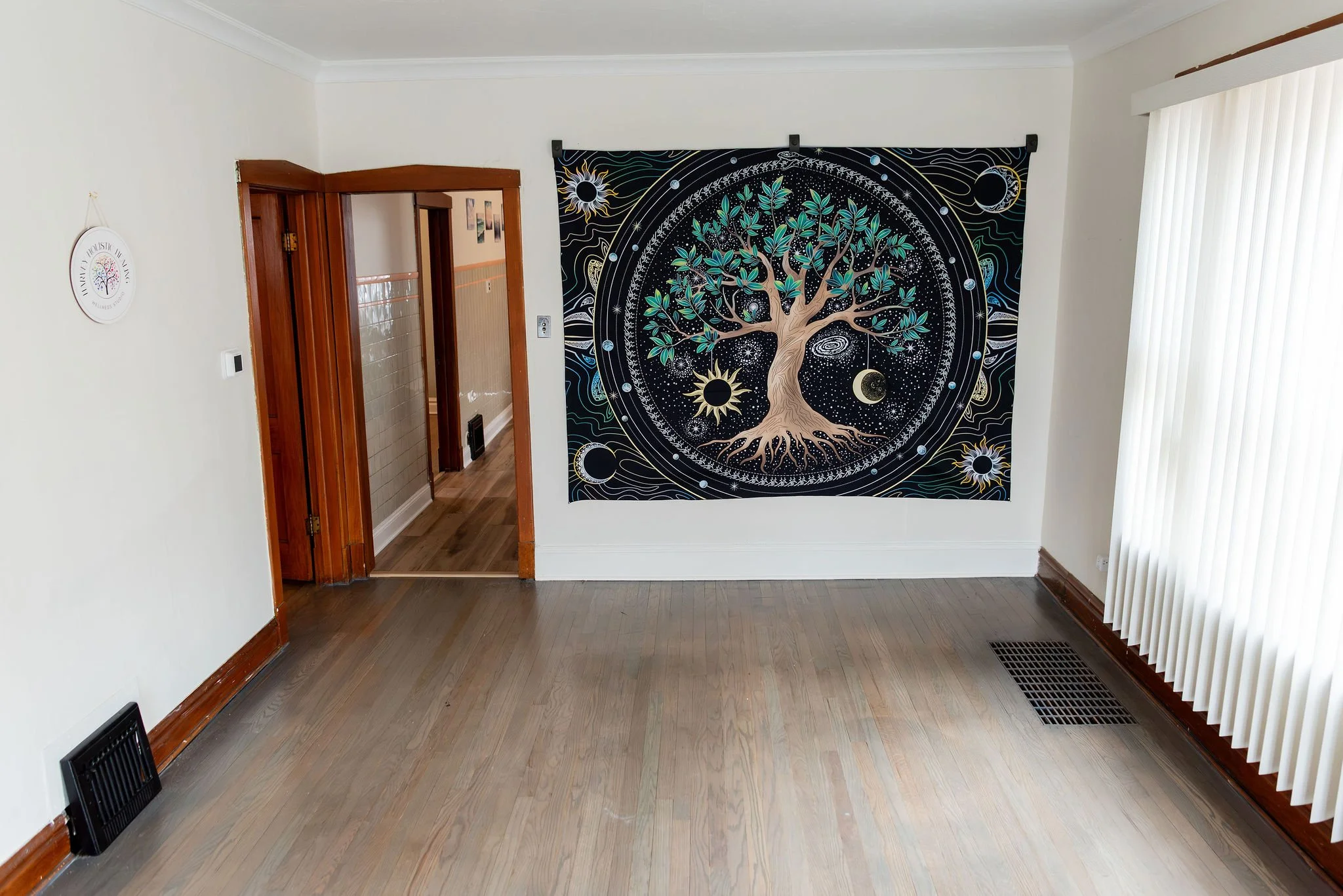 A room with light-colored walls, hardwood flooring, a large tapestry featuring a tree with celestial and cosmic designs, a sliding vertical blind window, and a doorway leading to a tiled area.