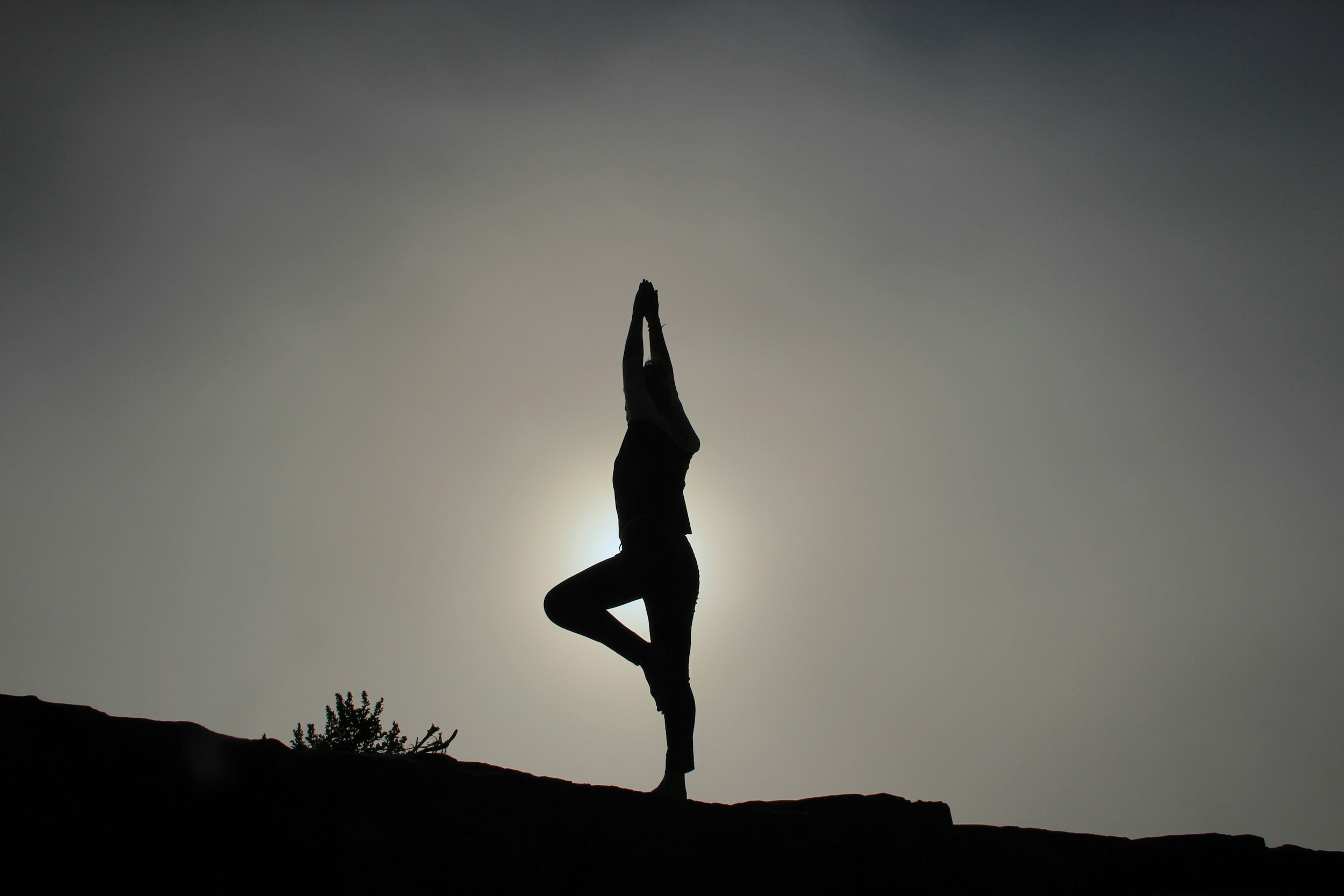 Person practicing yoga in a tree pose silhouette against the sun and sky.