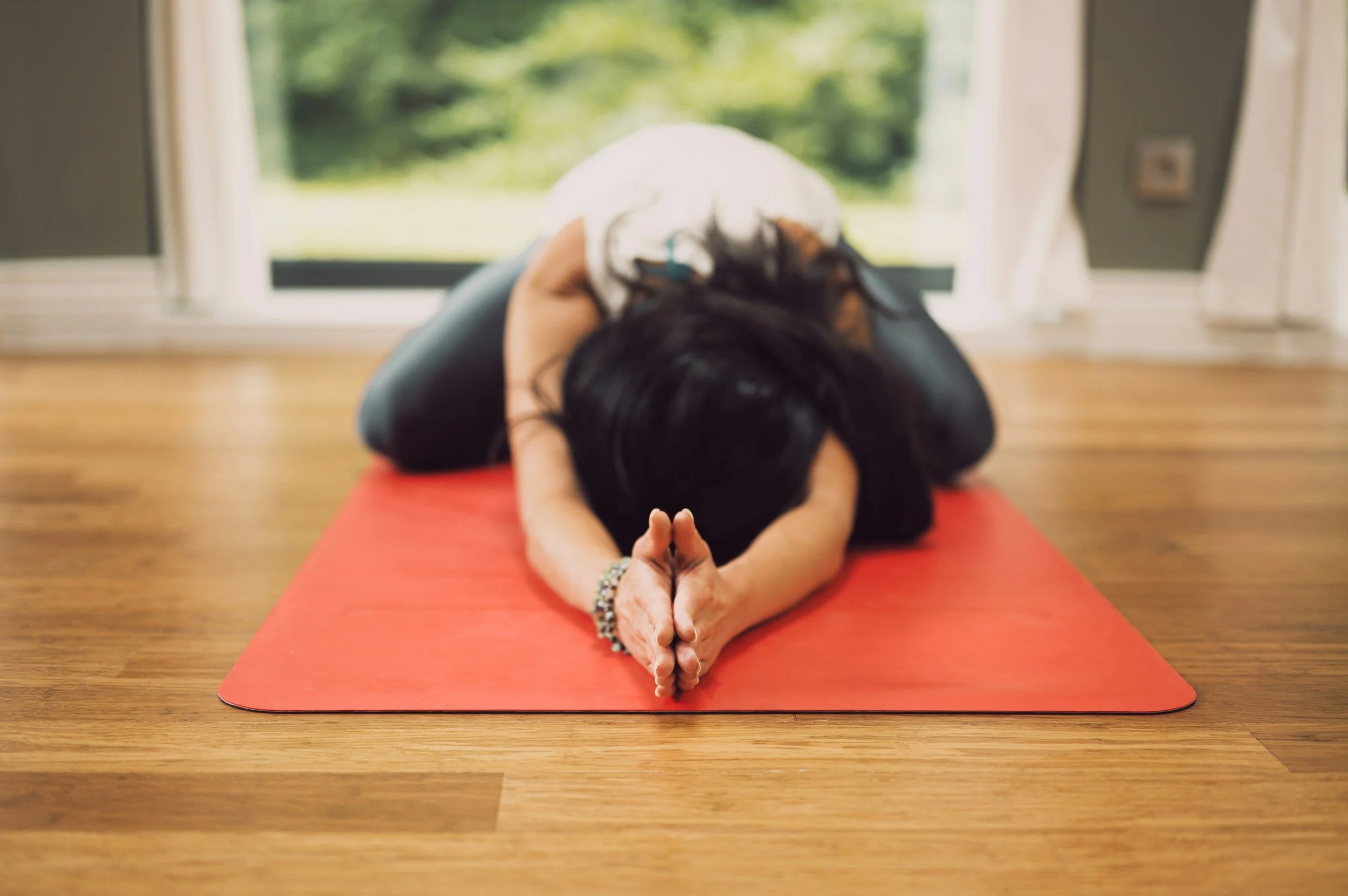 A woman practicing yoga indoors on a red mat, in a child's pose position with hands pressed together in front of her forehead, near a large window with greenery outside.