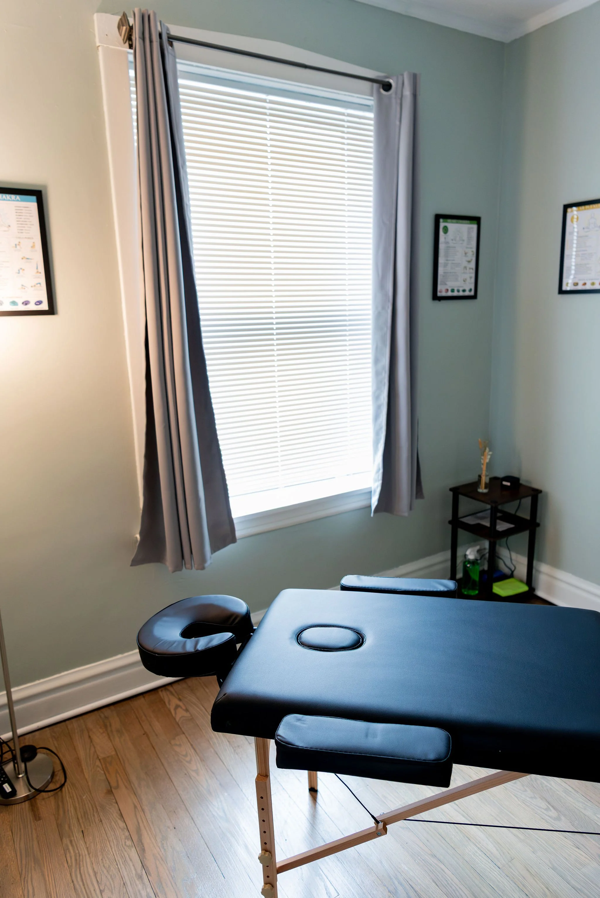 A massage table with a black cushioned surface and wooden frame, placed in a room with hardwood flooring, adjacent to a window with closed white blinds and light gray curtains, and small framed posters on the walls.
