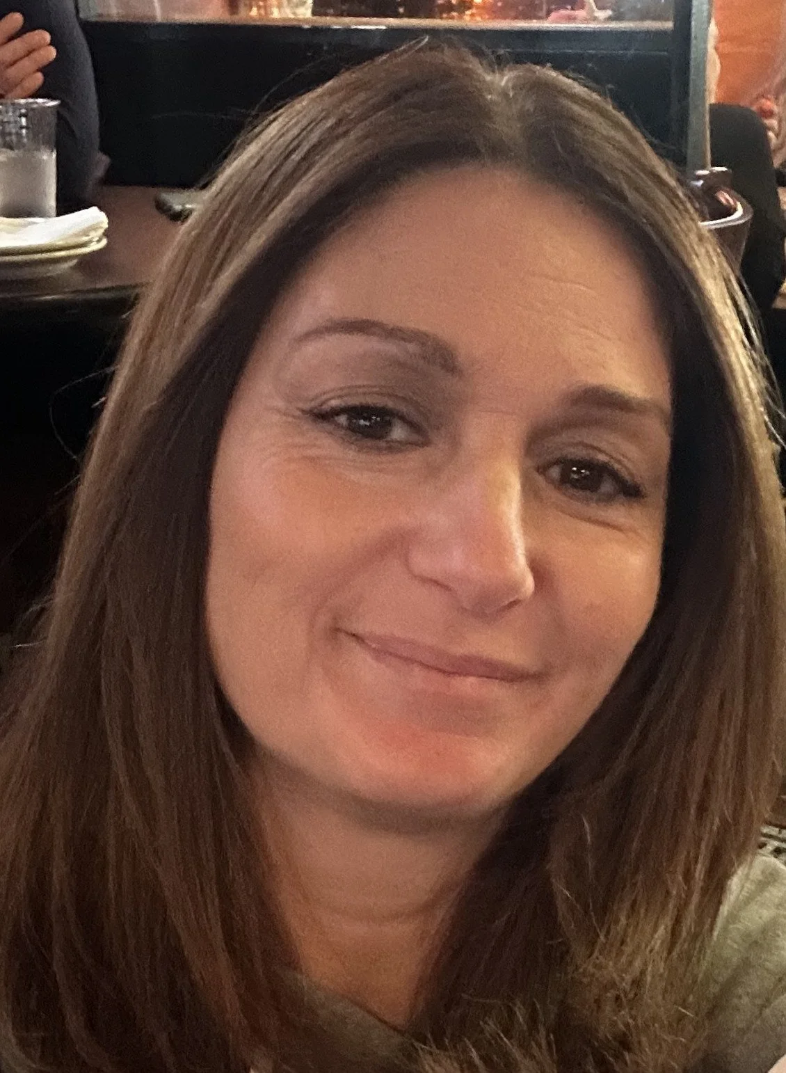 A woman with long brown hair wearing a sleeveless gray top, smiling softly in front of a white door.