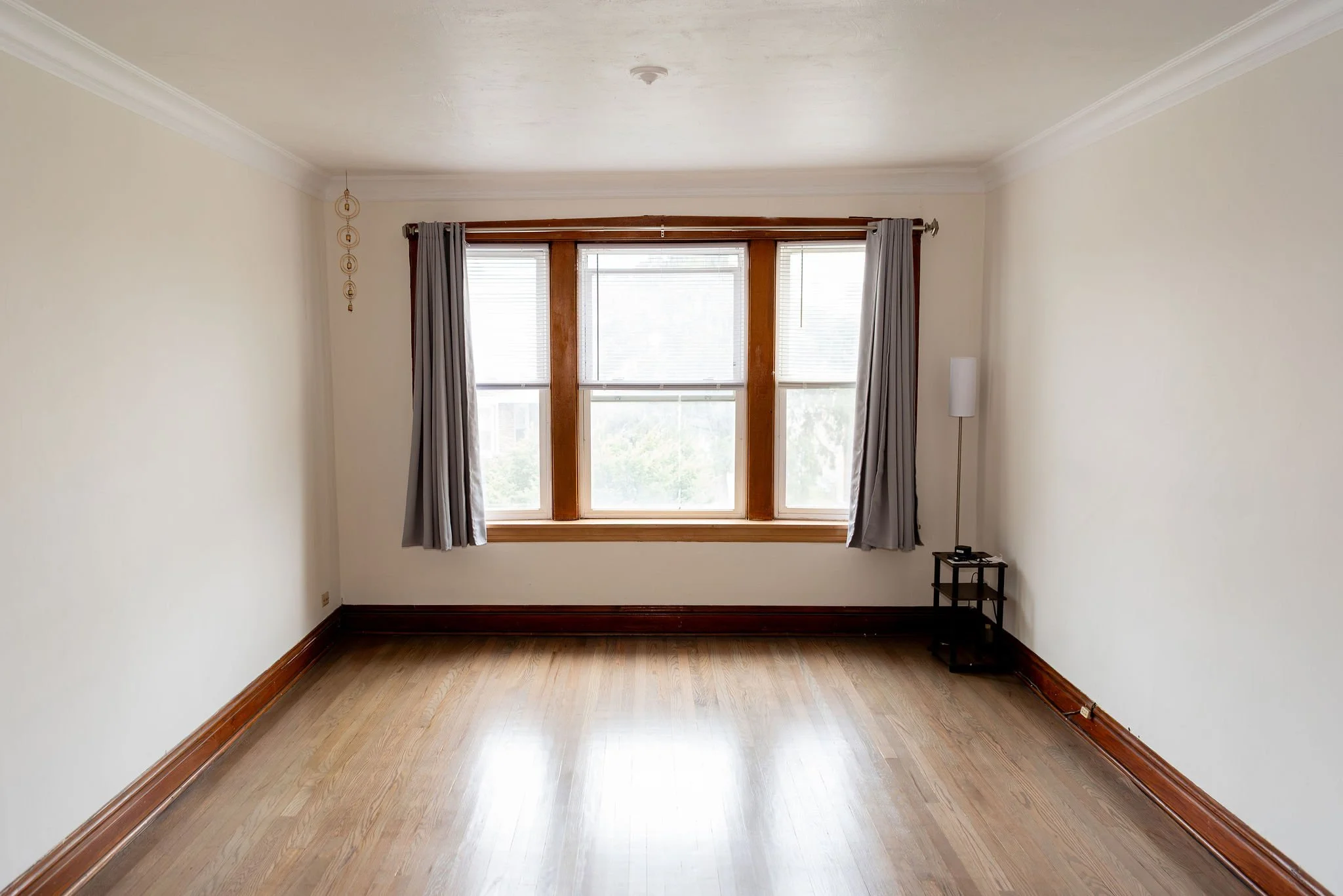 Empty room with hardwood floors, white walls, wooden window frame with three windows, grey curtains, and a small black table with phone and other items on it.