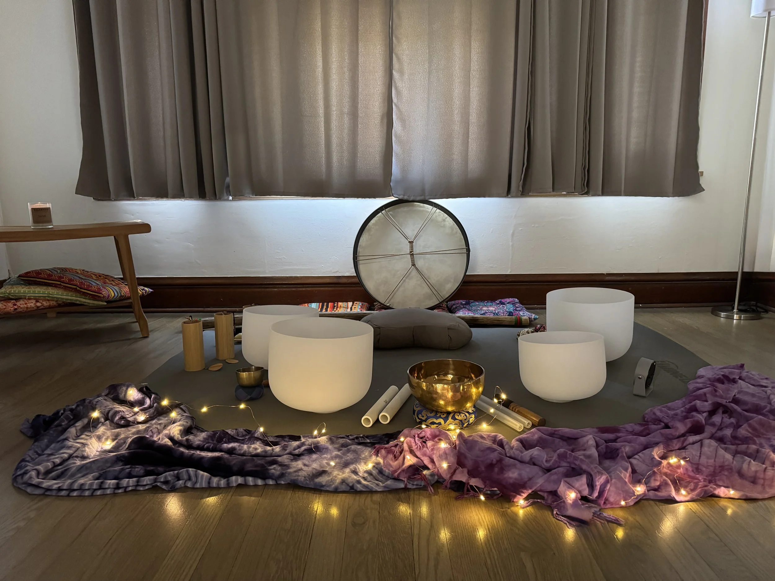 An indoor scene arranged for meditation or yoga, featuring a purple and black tie-dye blanket on the floor, surrounded by string lights, white crystal singing bowls, a brass bowl, rolled white paper or fabric, and colorful fabric cushions. In the background, a window with closed beige curtains is visible, and part of a wooden bench or table with a small brown object on it can be seen.