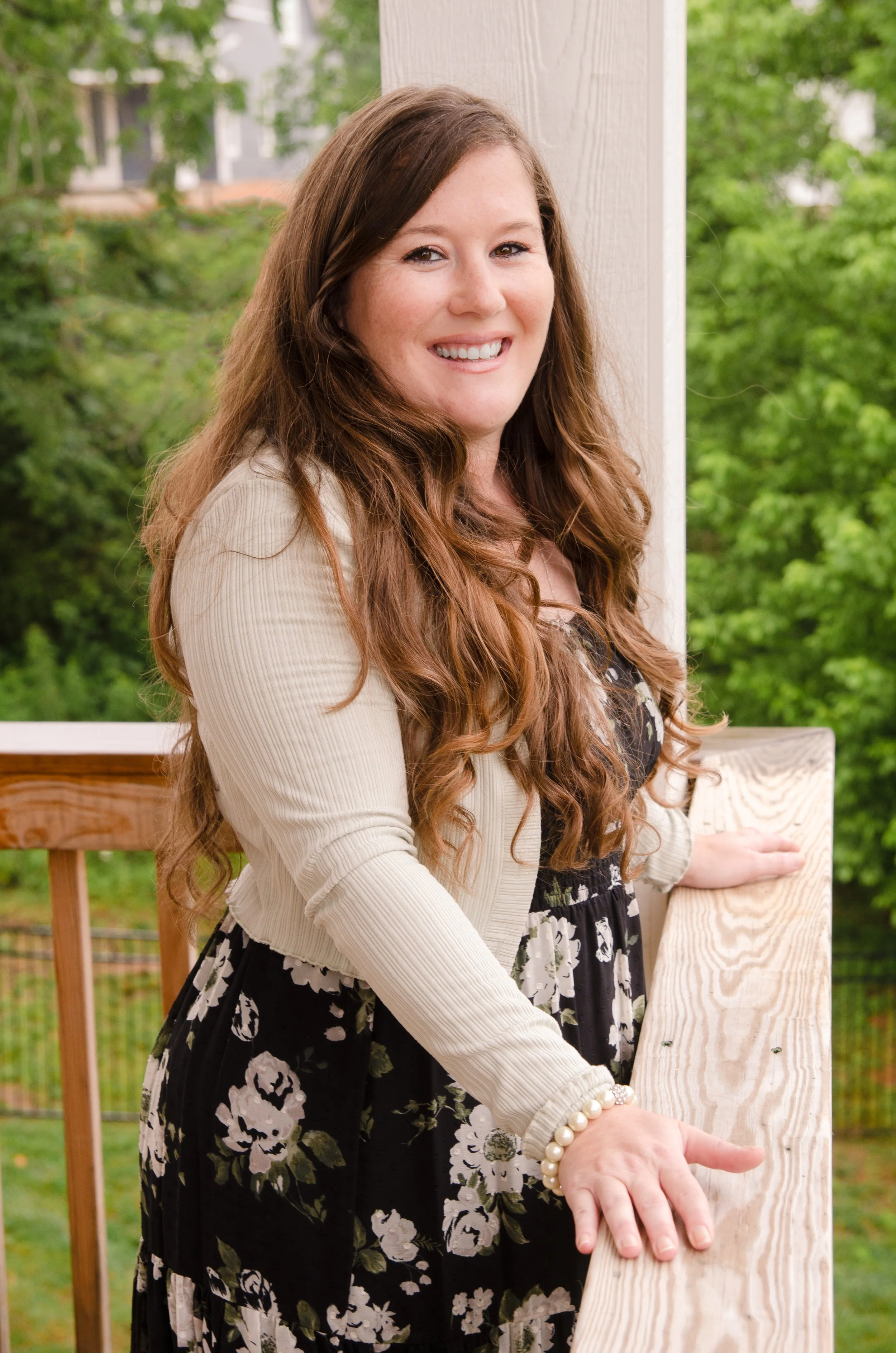 A woman with long, wavy brown hair smiling and standing outdoors on a balcony with a wooden railing and green trees in the background.