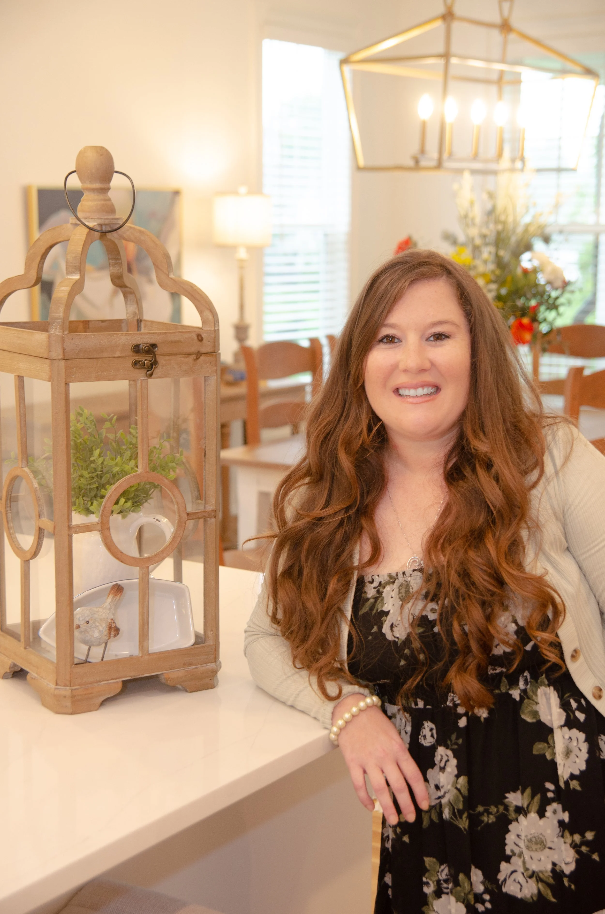 A woman with long red hair wearing a floral dress and light-colored cardigan, smiling and leaning on a white kitchen countertop next to a decorative lantern with greenery and a small bird figurine inside, in a well-lit dining area.