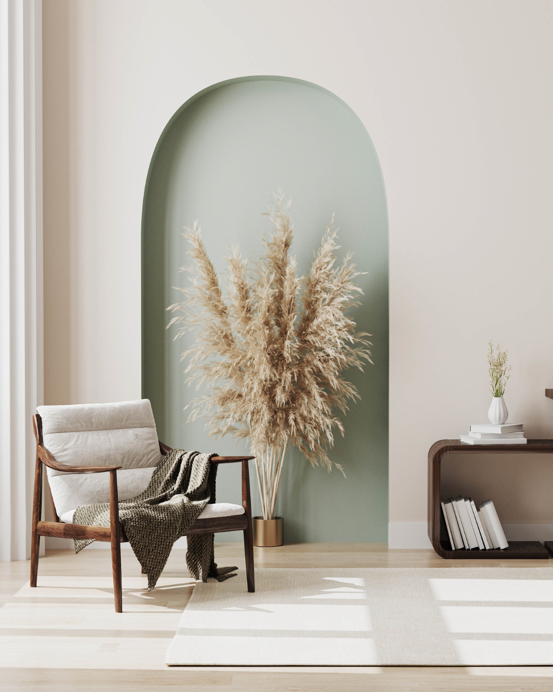 Modern interior with a wooden armchair draped with a blanket, a large beige pampas grass arrangement in a gold planter, a small bookshelf with a white vase and books, and a green arched wall accent.