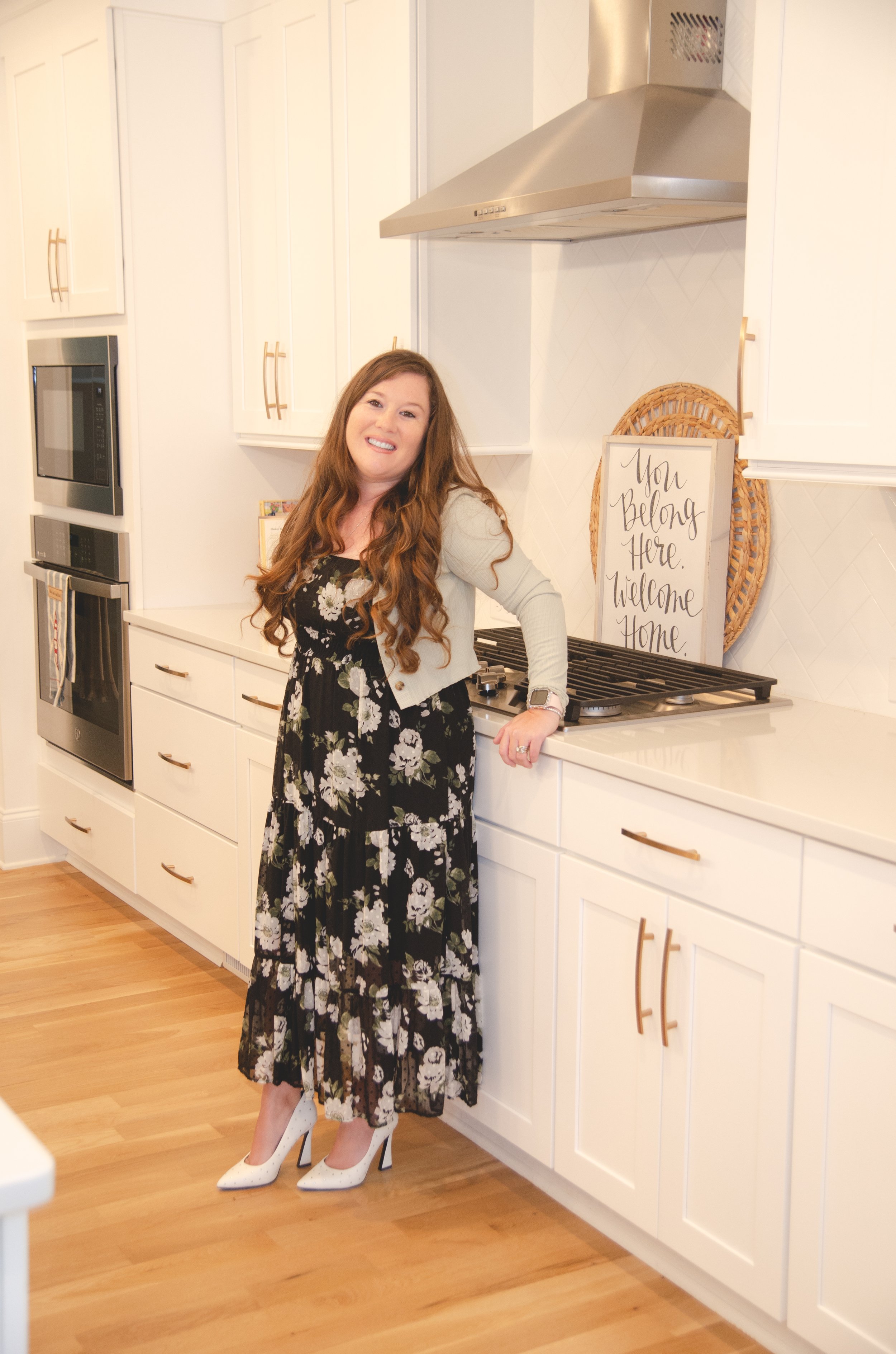 A woman with long red hair wearing a floral black dress, white cardigan, and white high heels standing in a bright white kitchen with wooden floors, smiling at the camera.