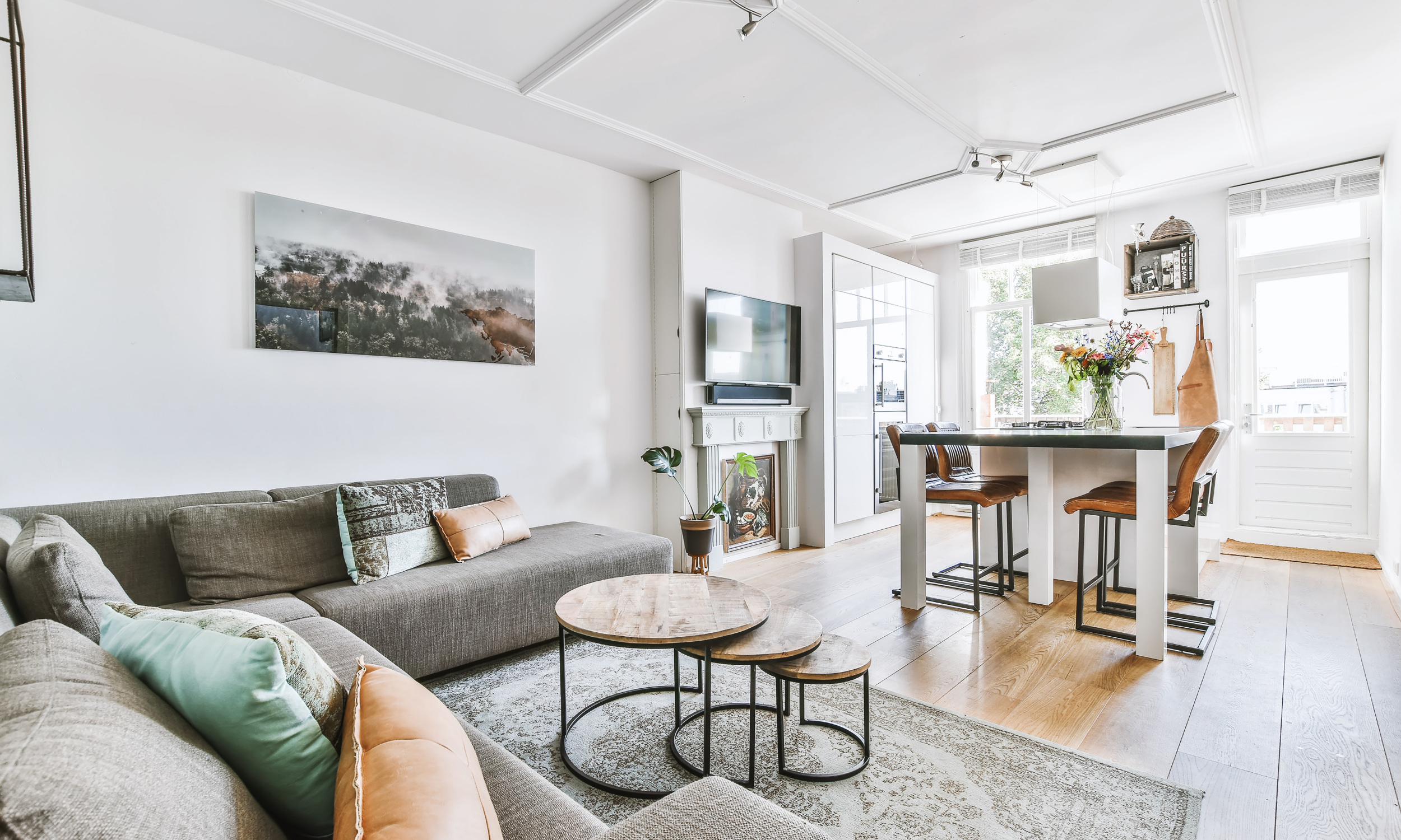 A bright, modern living room with a gray sofa, a set of nested round wooden coffee tables, a wall-mounted TV above a white fireplace, and a kitchen area with a white dining table and brown chairs near large windows with outdoor views.