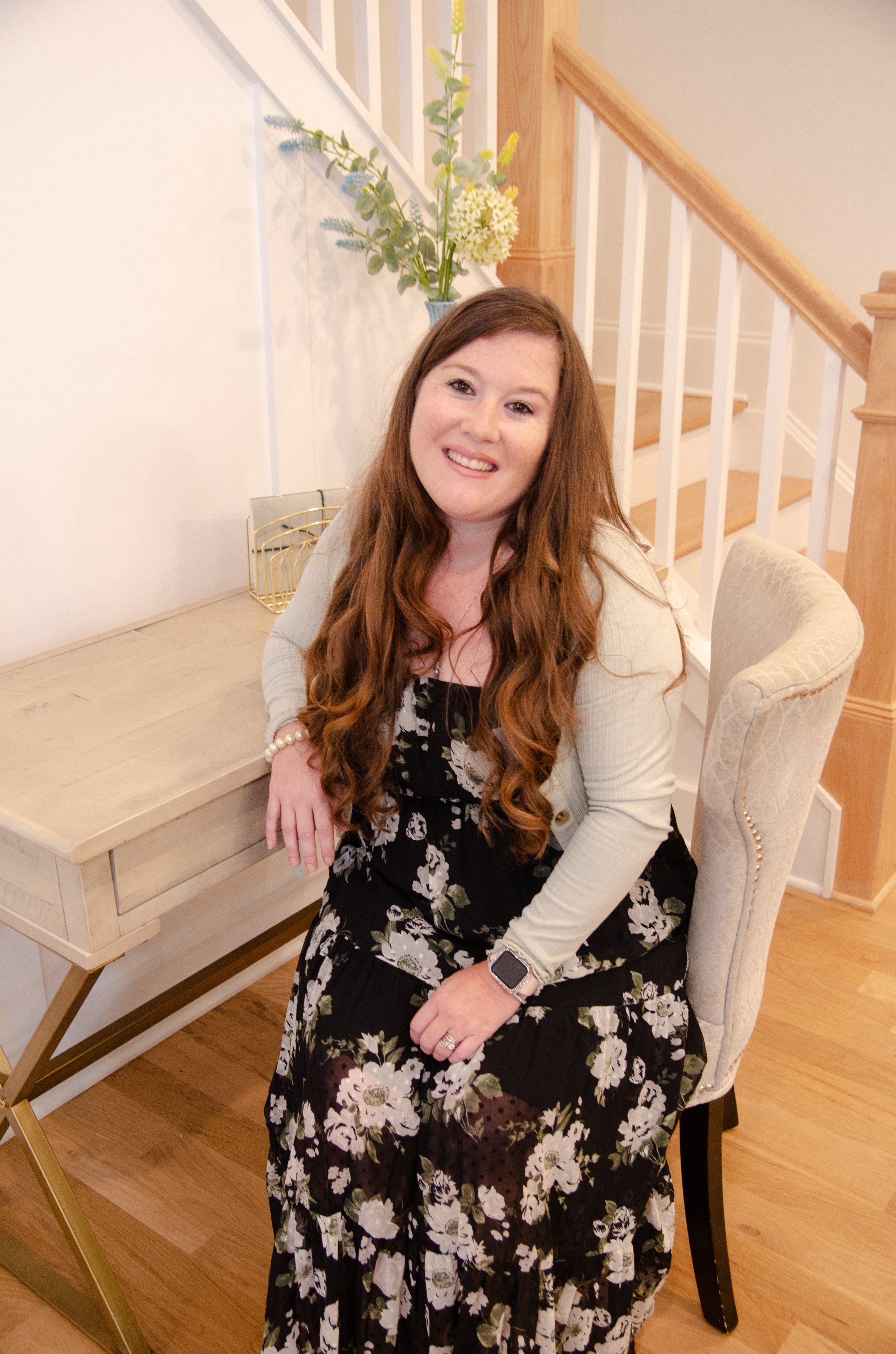 A woman with long red hair, wearing a black floral dress and a light-colored cardigan, sits on a beige upholstered chair at a wooden desk in a home interior with stairs in the background.