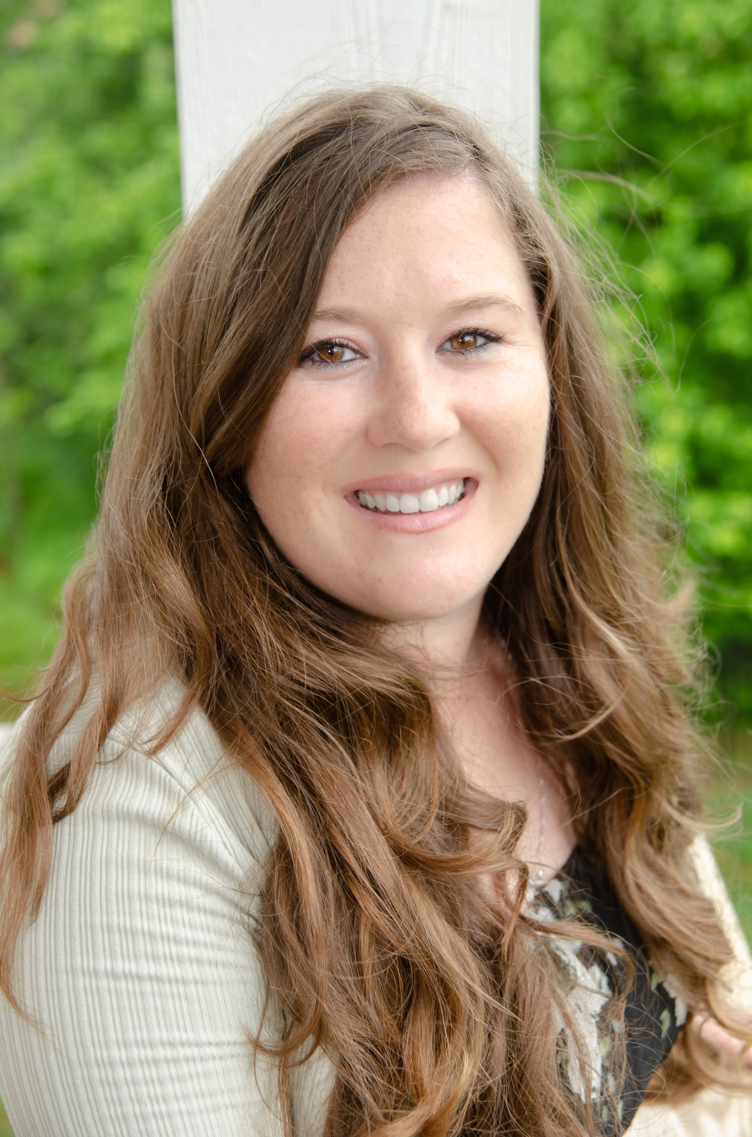 A young woman with long, wavy brown hair smiling outdoors, with green foliage in the background.