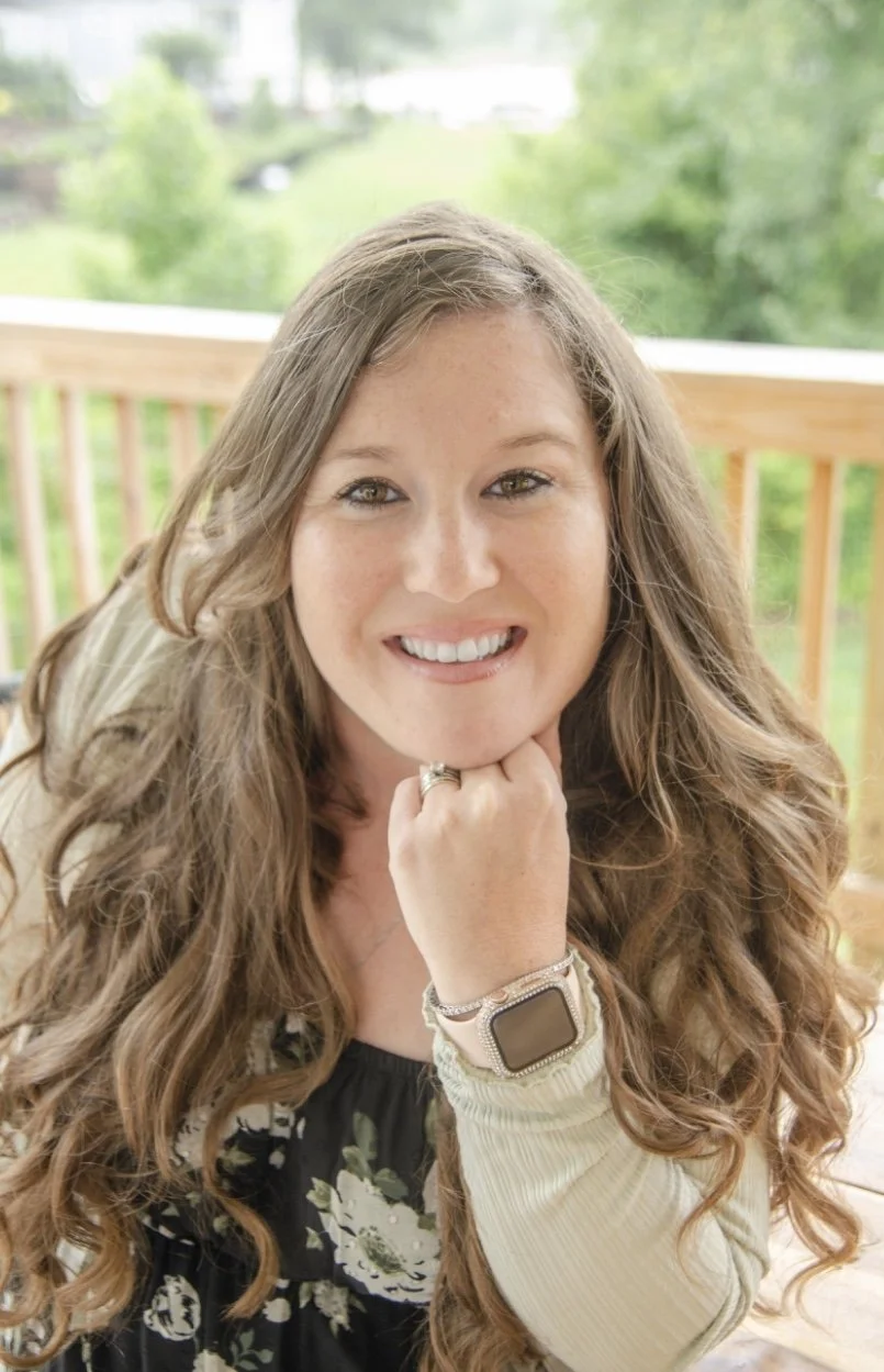 A woman with long, wavy hair smiling and resting her chin on her hand, standing outdoors with a blurred green landscape background.