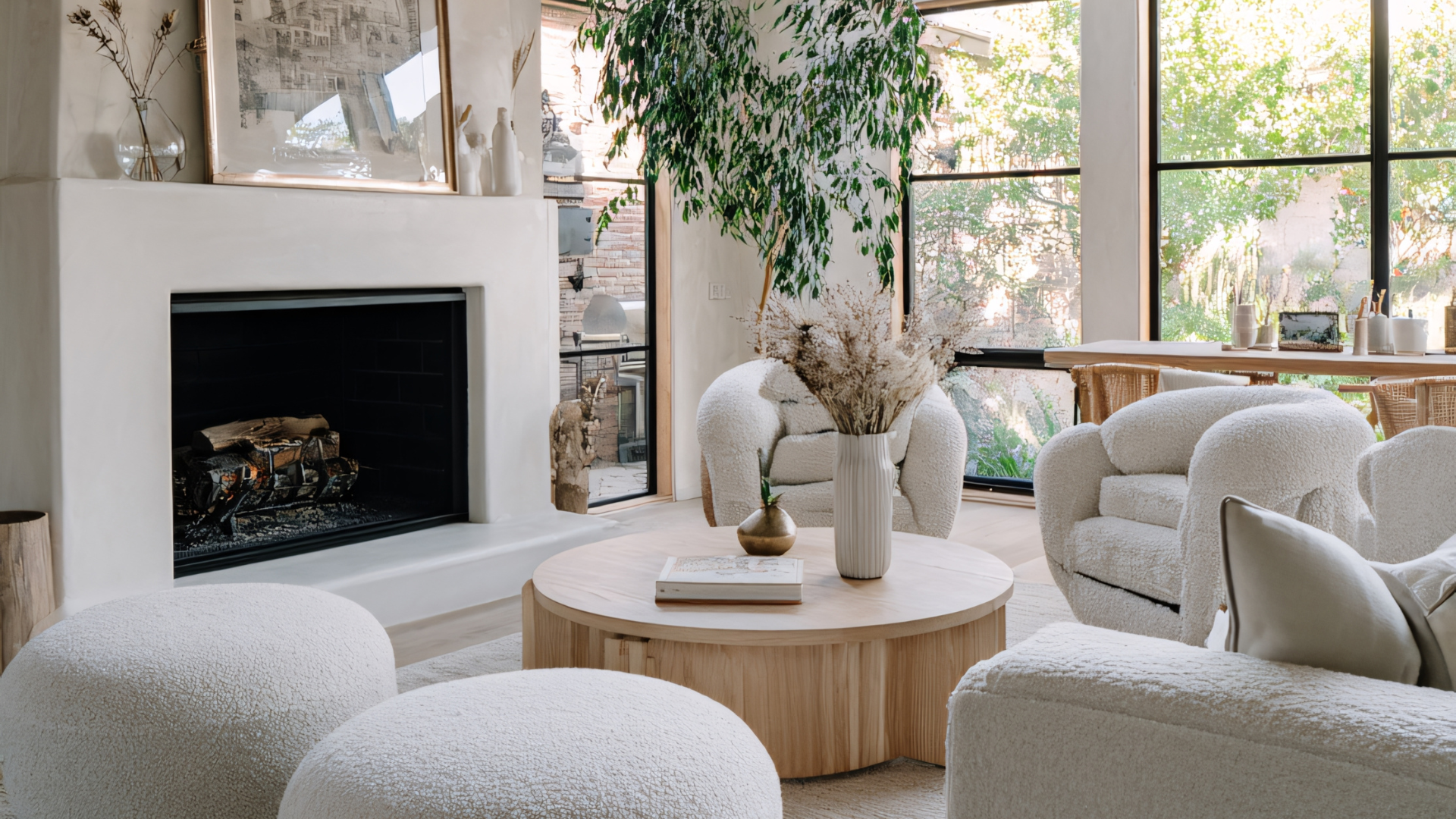 A bright living room with large windows, a white fireplace, and a round wooden coffee table with a vase of dried flowers and a book. There are white, textured armchairs and ottomans, a big green plant, and a dining table with chairs in the background.