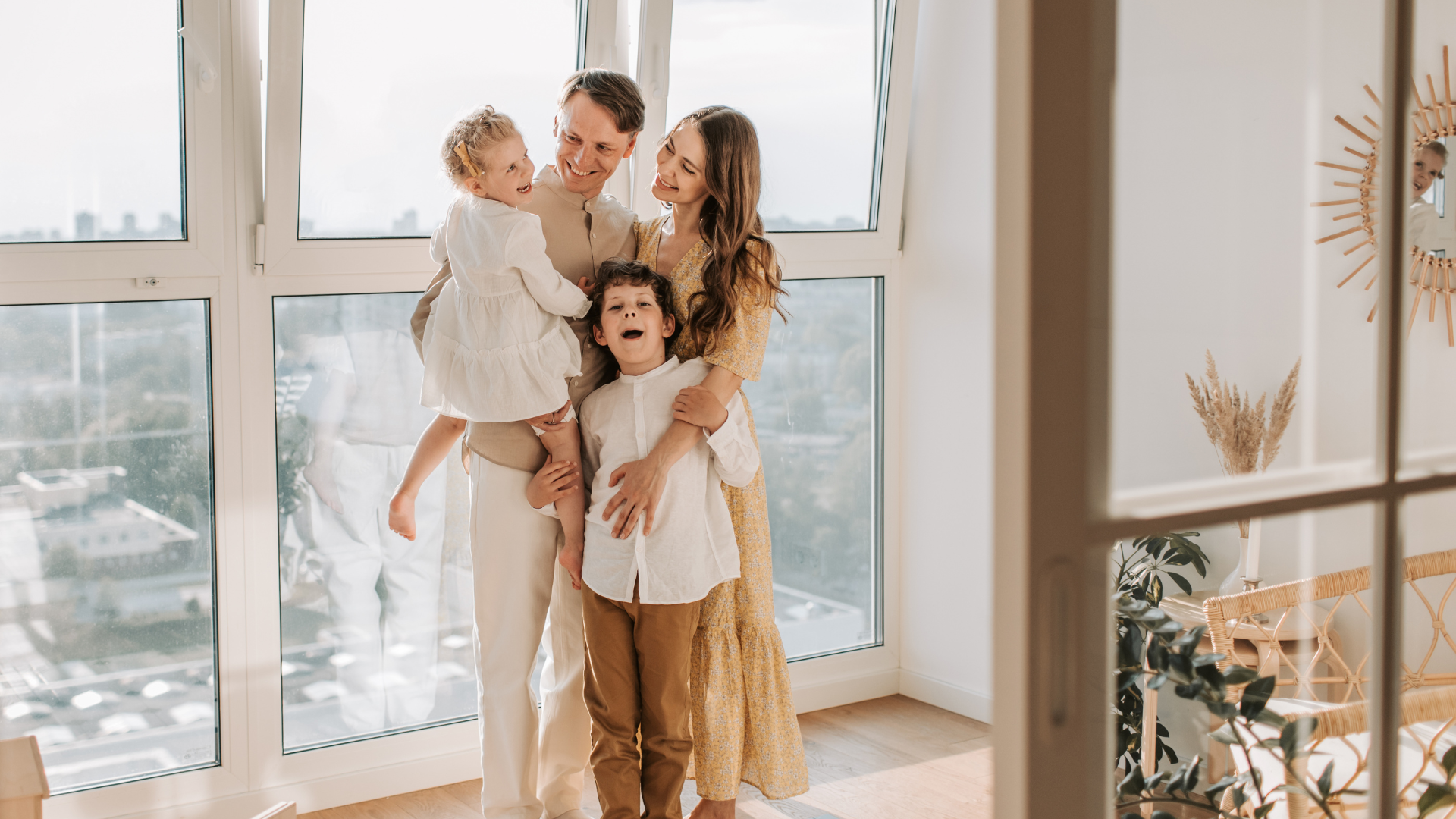 A happy family of four stands together near large windows in a sunlight-filled room, smiling and enjoying each other's company.