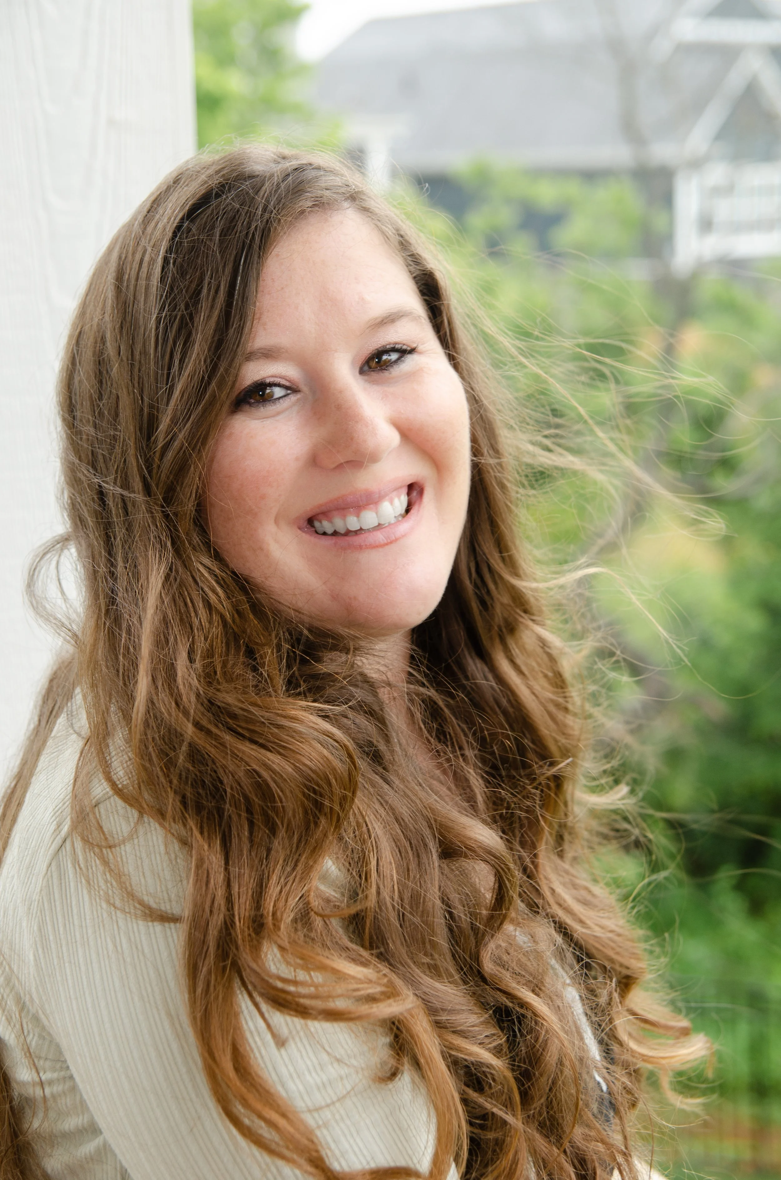 A young woman with long, wavy, brown hair, smiling and leaning against a window with a green, outdoor background.