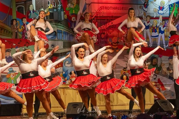 Tanzgruppe in roten und weißen Kostümen auf einer Bühne mit bunter Kulisse, performend in einer festlichen Manege.