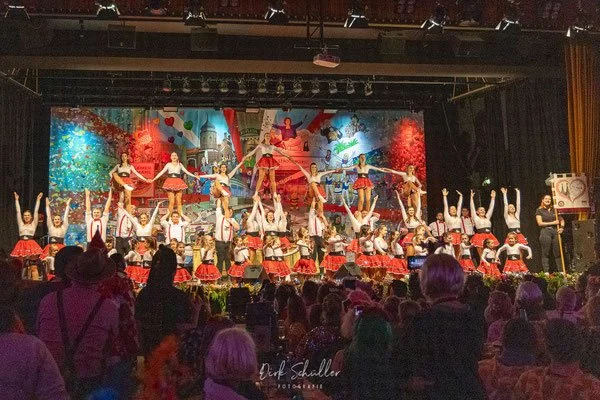 Tanzgruppe auf einer Bühne bei einer Aufführung, mit einem bunten Banner im Hintergrund, ein Publikum sitzt vor der Bühne.