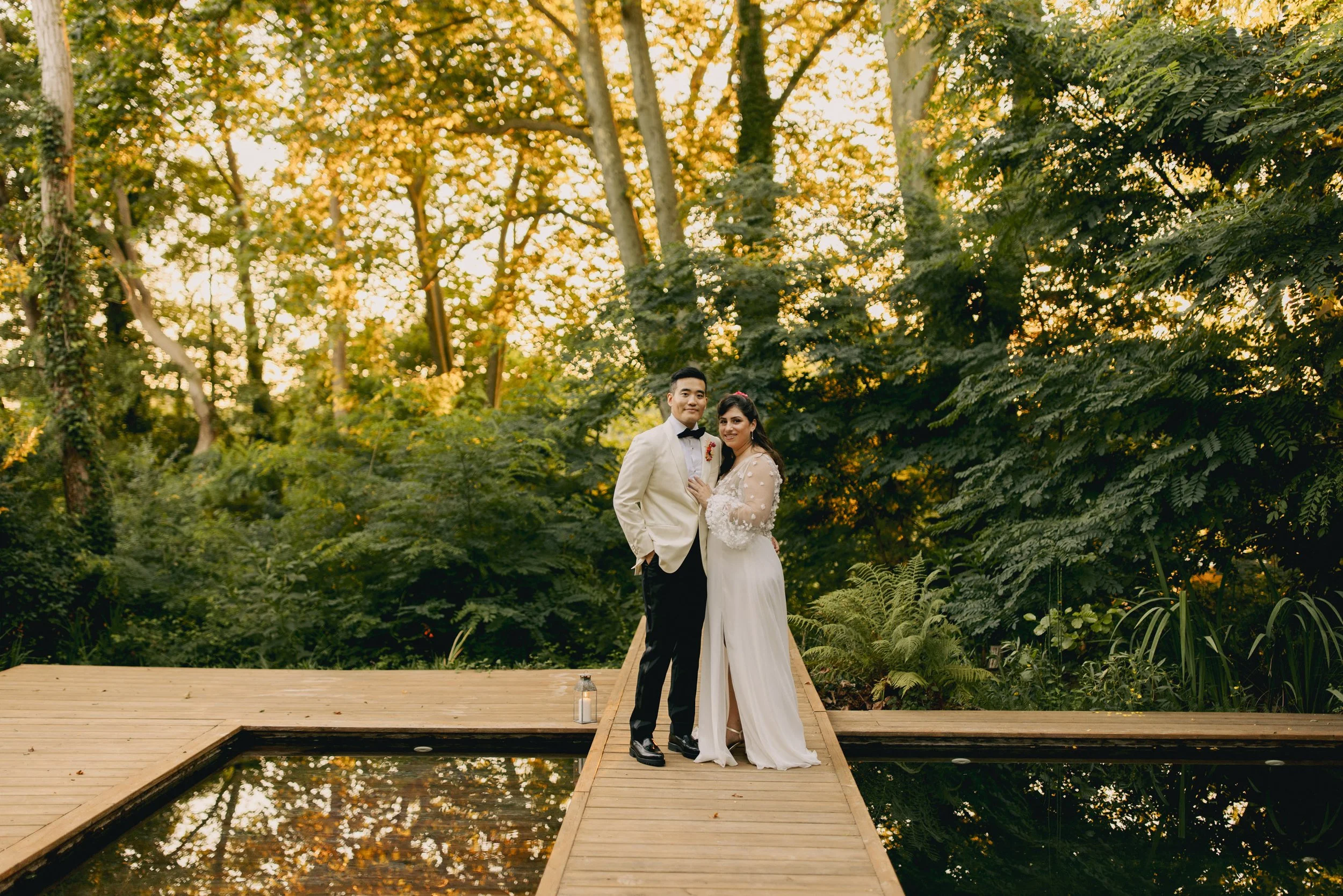 Un couple en mariage posant sur une passerelle en bois entourée de verdure avec arbres, lors d'un coucher de soleil.