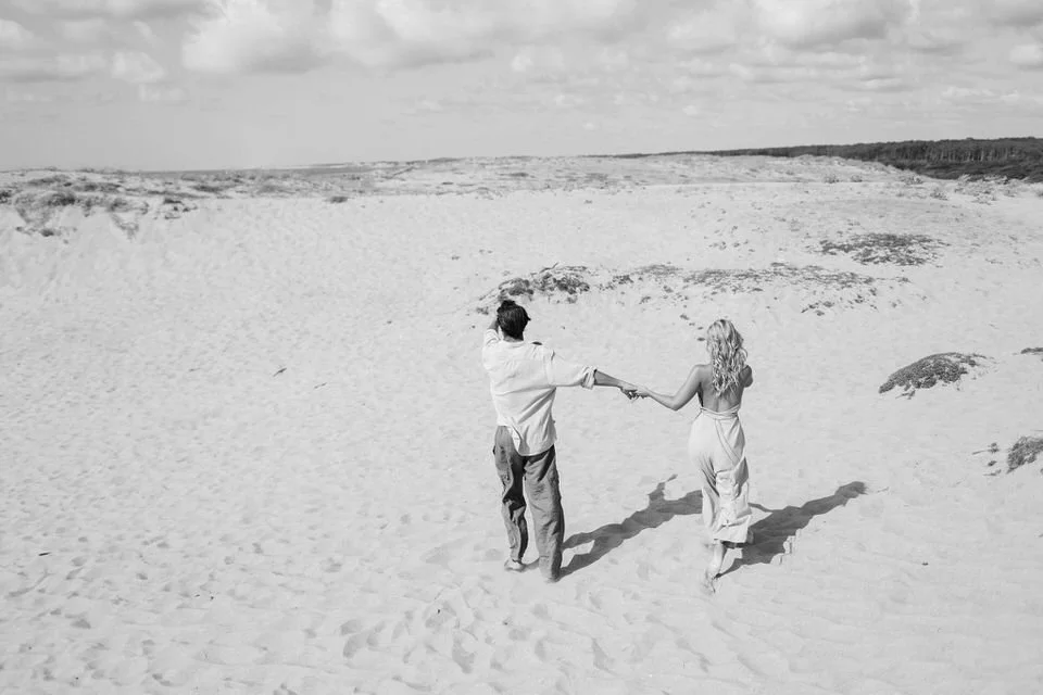 Un couple marche sur la plage en se tenant la main, vue de dos, dans une scène en noir et blanc.