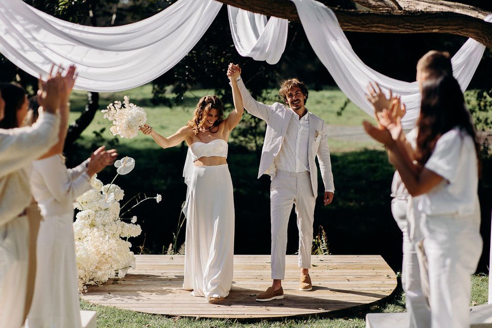 Une mariée et un marié souriants tenant la main levée, se tenant sur une scène en bois décorée avec des draperies blanches, entourés par des invités qui applaudissent, lors d'un mariage en plein air