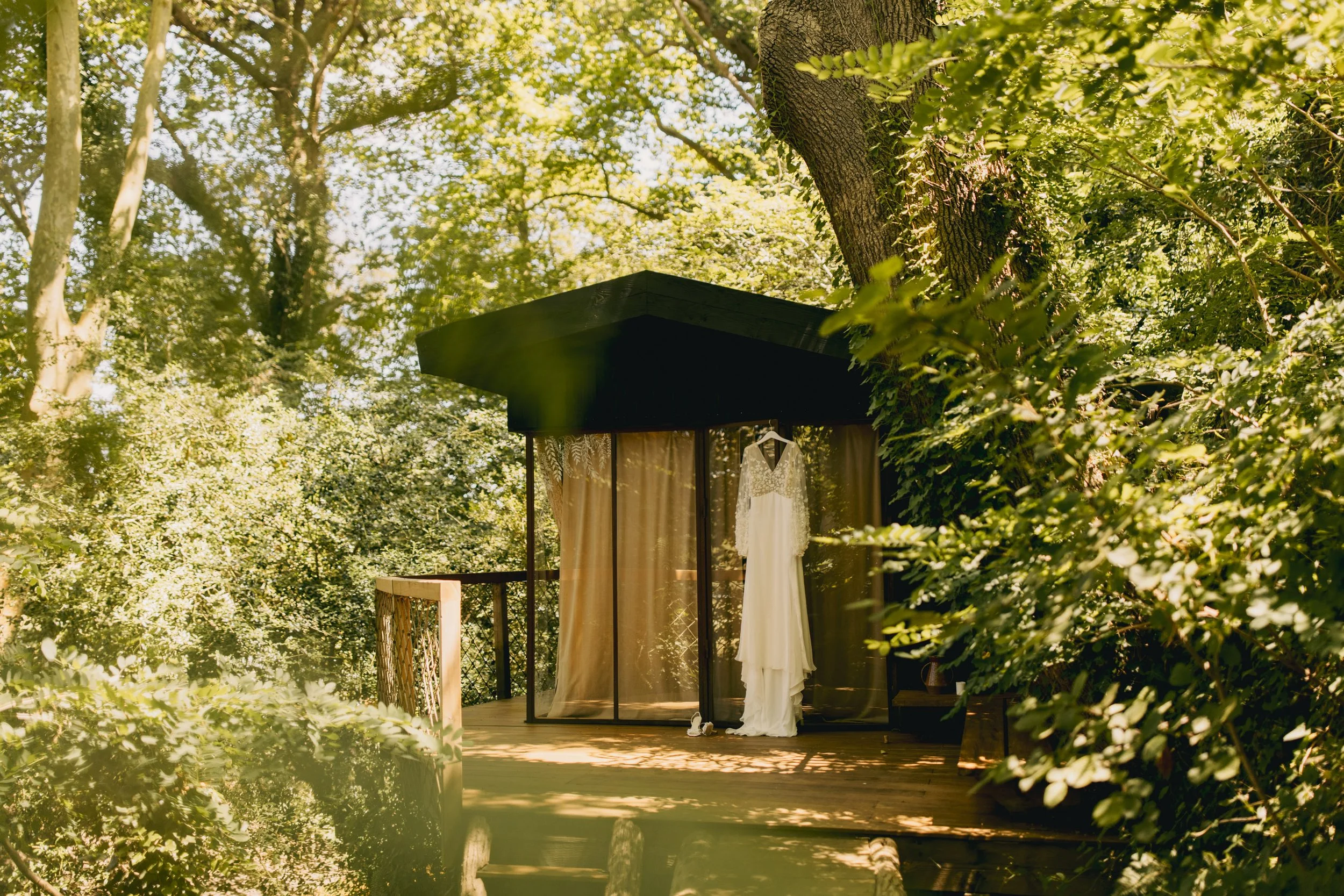 Une robe de mariée blanche suspendue sur un support à l'intérieur d'une cabane en bois entourée d'arbres verts. La scène est baignée de lumière naturelle dans un environnement forestier.