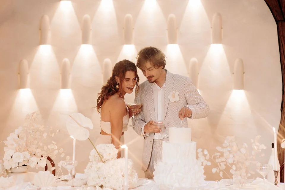 Un couple en vêtements de mariage célébrant leur mariage avec un gâteau, entouré de fleurs blanches, dans une décoration chaleureuse