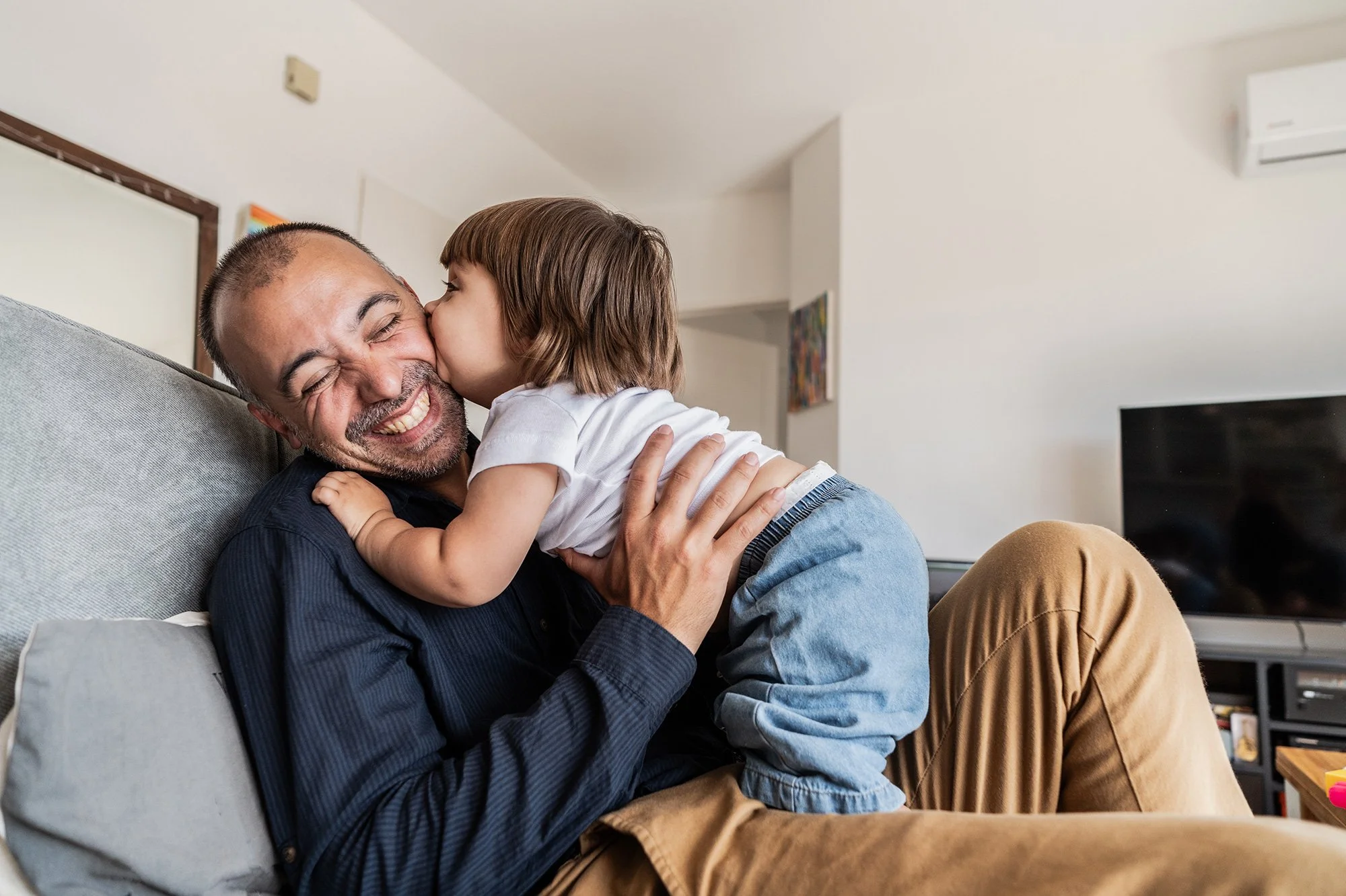 A young girl giving a kiss to a smiling man on the face while sitting on a couch in a living room.