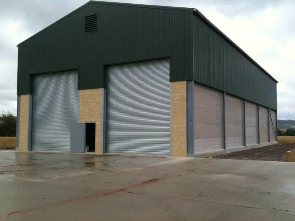 A large commercial storage building with three gray roll-up garage doors and a small pedestrian door, made of brick and metal siding, on a paved surface.