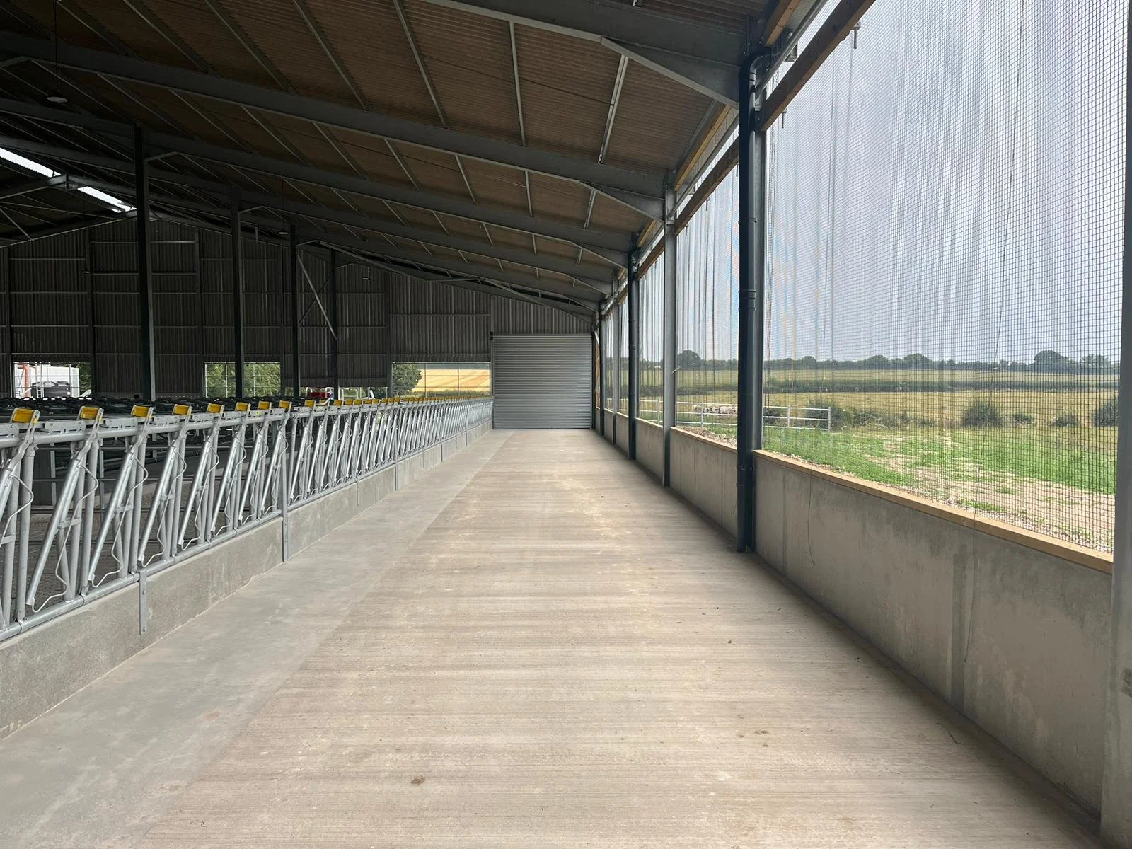 Empty covered walkway with metal guardrail on the left and a wire mesh fence on the right, overlooking a farm field.
