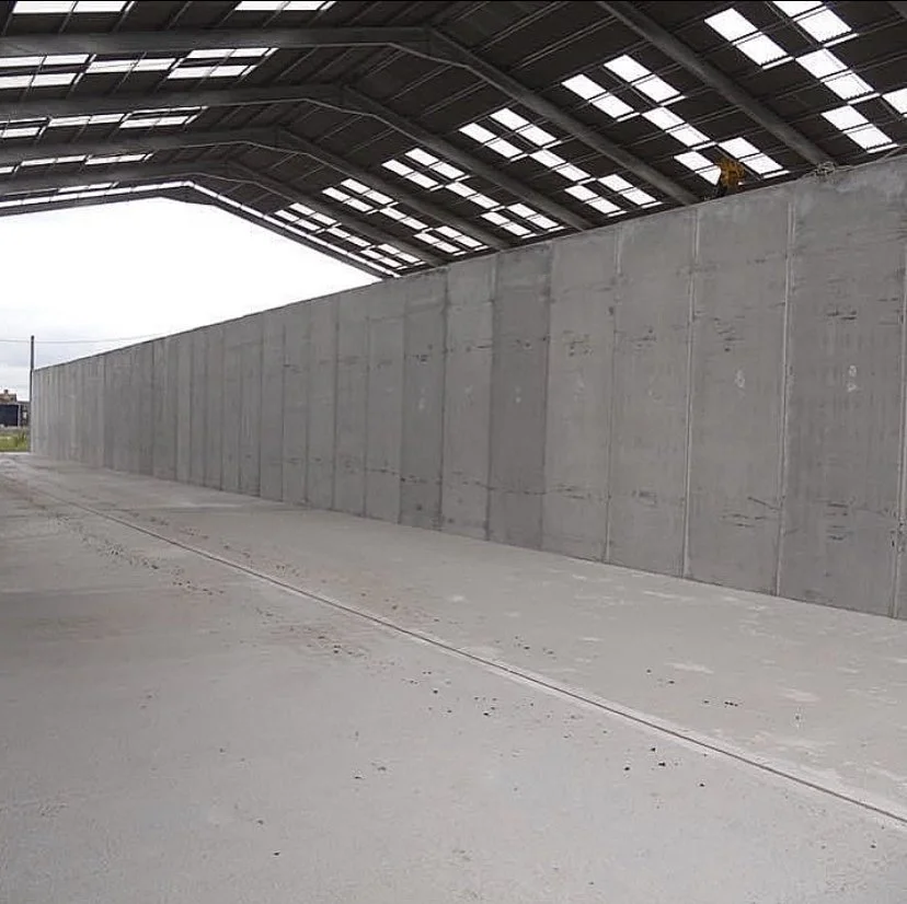 A concrete driveway runs alongside a tall concrete wall that separates an empty parking or loading area from an indoor structure with a high, sloped roof made of metal panels with skylights.