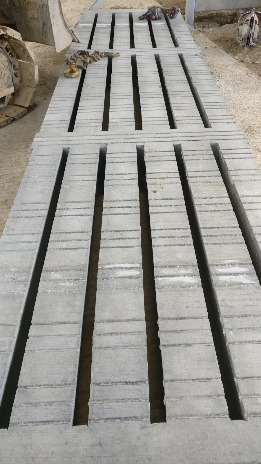 Long concrete blocks with elongated slots, stacked on a work table at a construction site, with tools and equipment nearby.