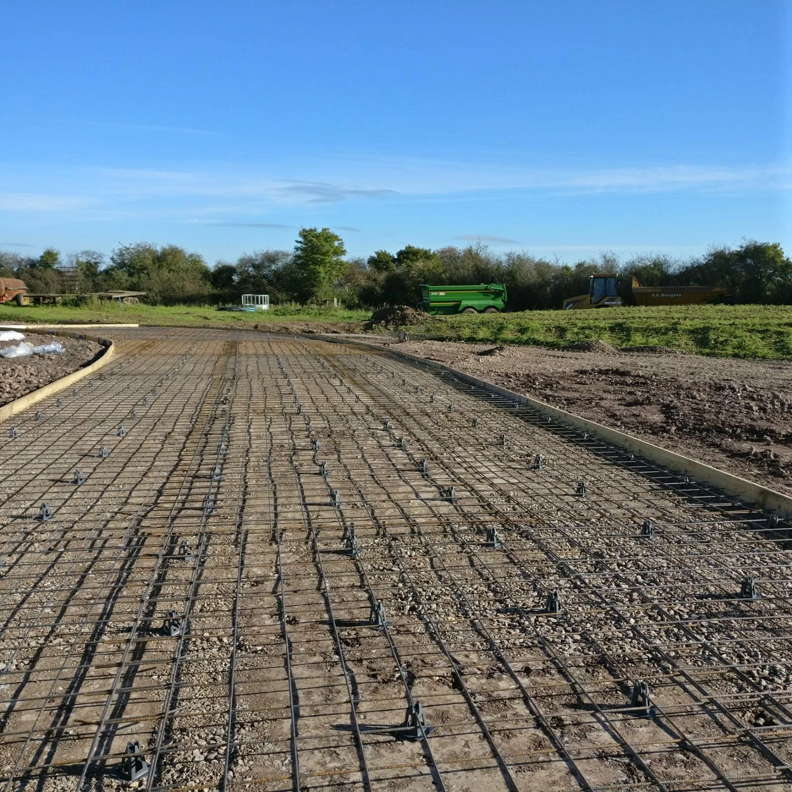Construction site with rebar laying on the ground, curved concrete border, and construction equipment like a green trailer and a yellow bulldozer in the background under a clear blue sky.
