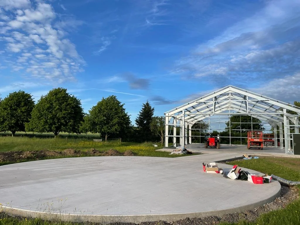 A greenhouse under construction with a curved concrete foundation in a rural area, surrounded by green trees and a blue sky with clouds.
