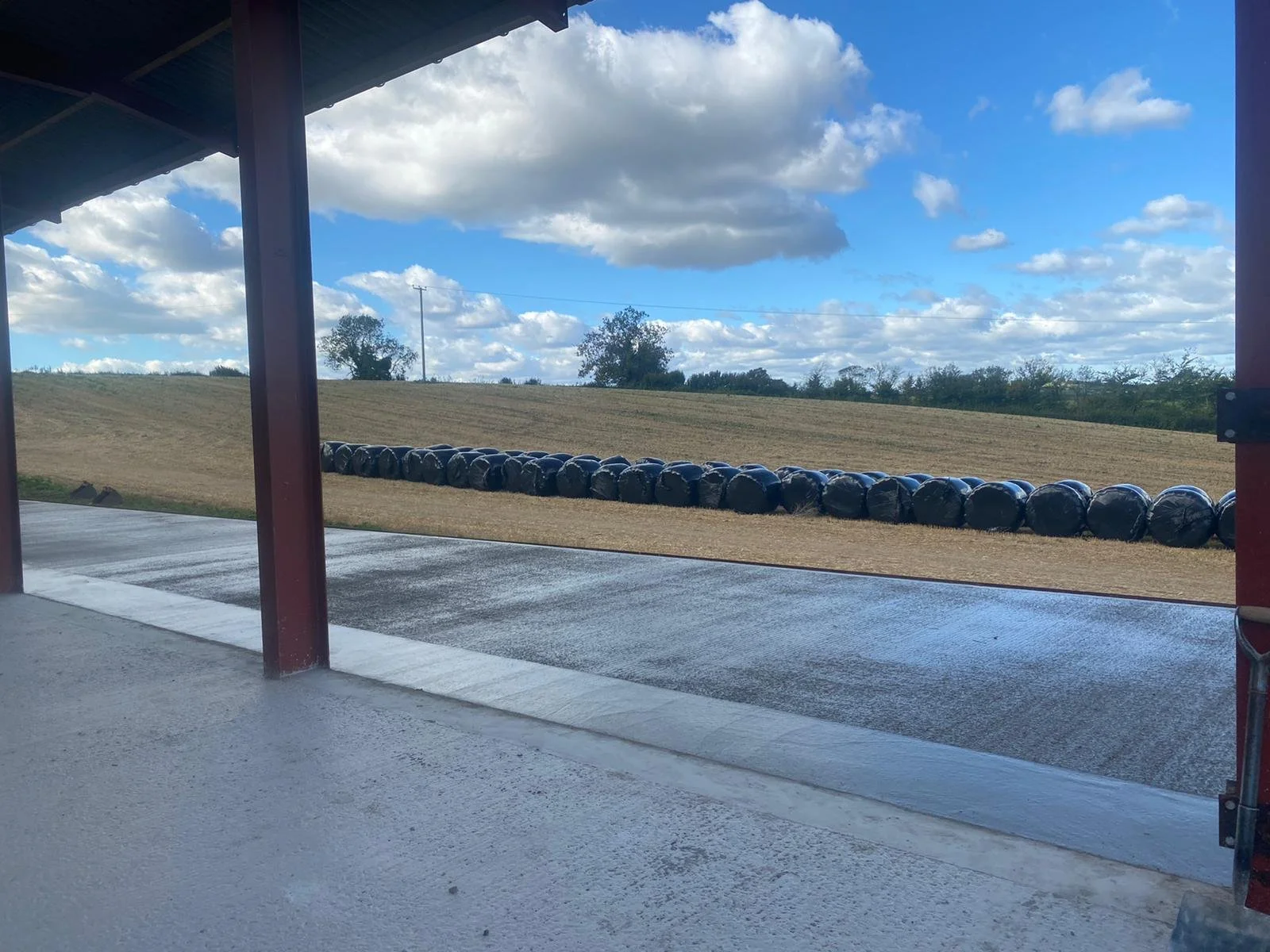 View of a farm field with hay bales wrapped in black plastic, seen from under a covered structure on a cloudy day.