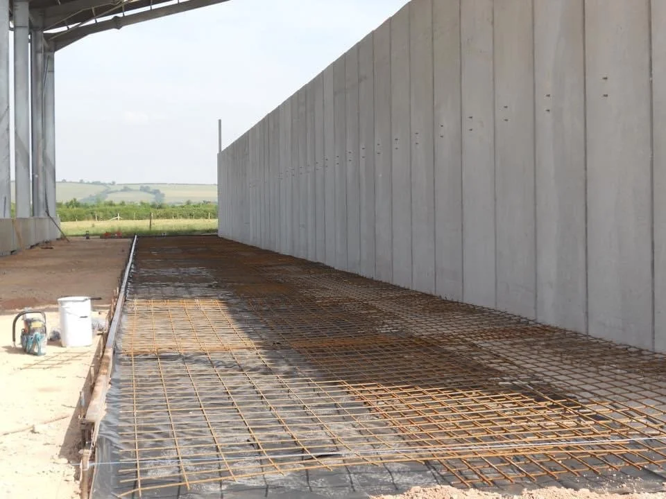 Construction site with rebar grid on ground inside a covered area, with a concrete wall on the right and open landscape in the background.