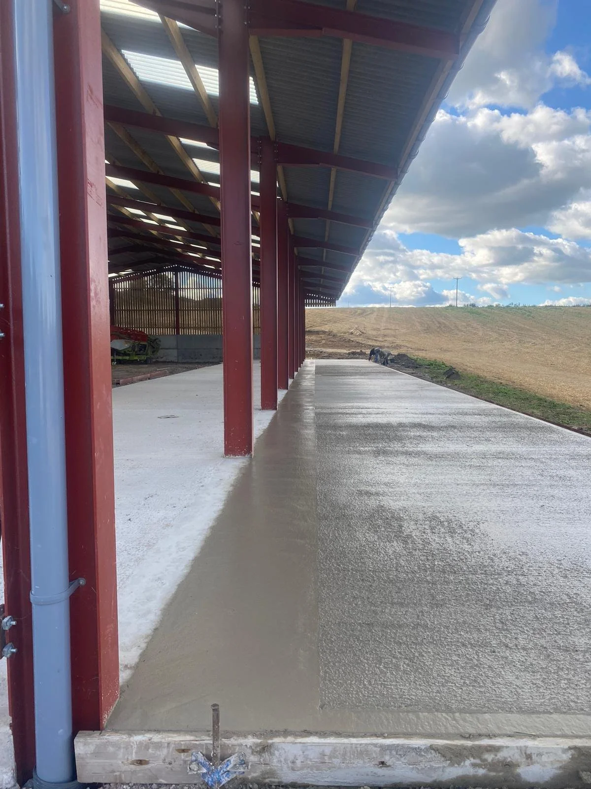 Concrete pathway under construction with a metal and wood roof structure, open landscape with a cloudy sky in the background.