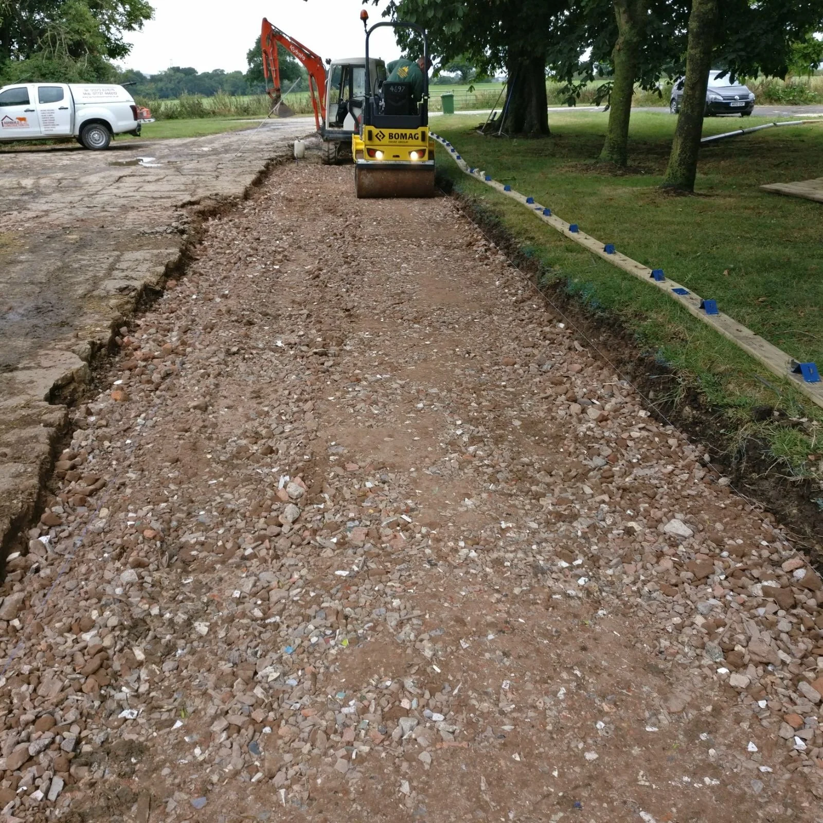 Construction site with a small excavator working on a gravel pathway, surrounded by trees and parked vehicles.