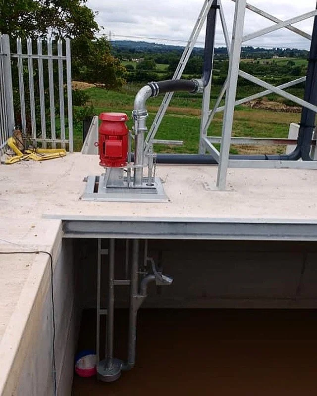 A rooftop with industrial piping, a red pump, and a metal safety railing, overlooking a rural landscape with fields and trees.
