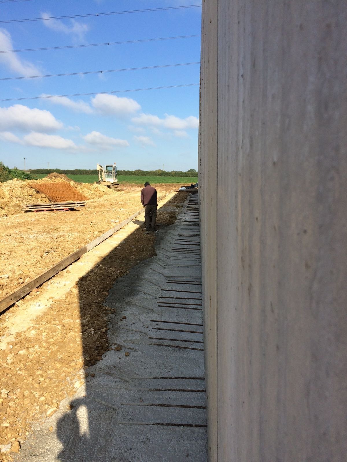 Construction site with a worker standing on gravel next to a concrete wall, with an excavator and sand piles in the background under a partly cloudy sky.
