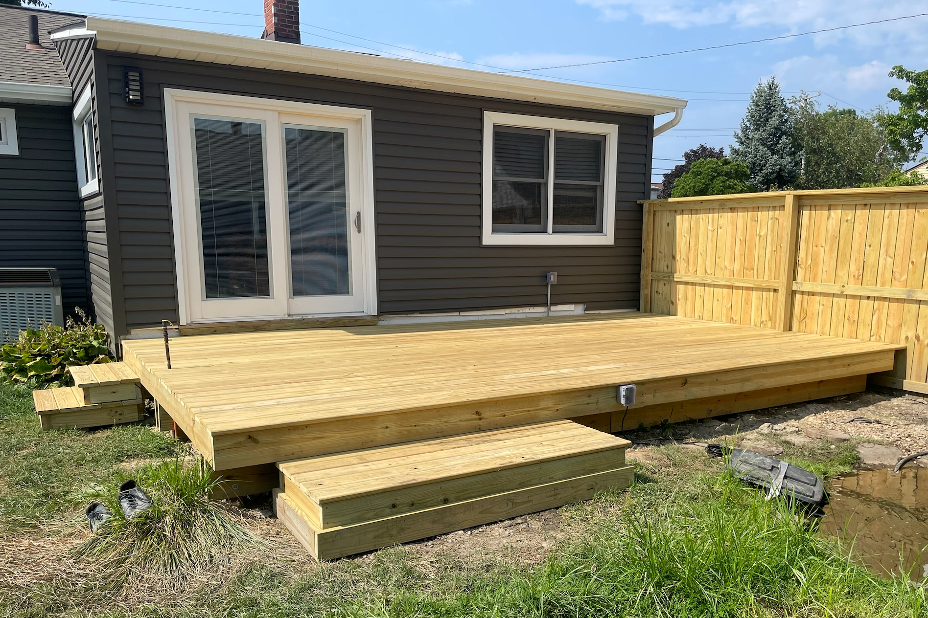 Newly built wooden deck attached to a house with sliding glass doors and a window, surrounded by a wooden fence and some green grass and plants.