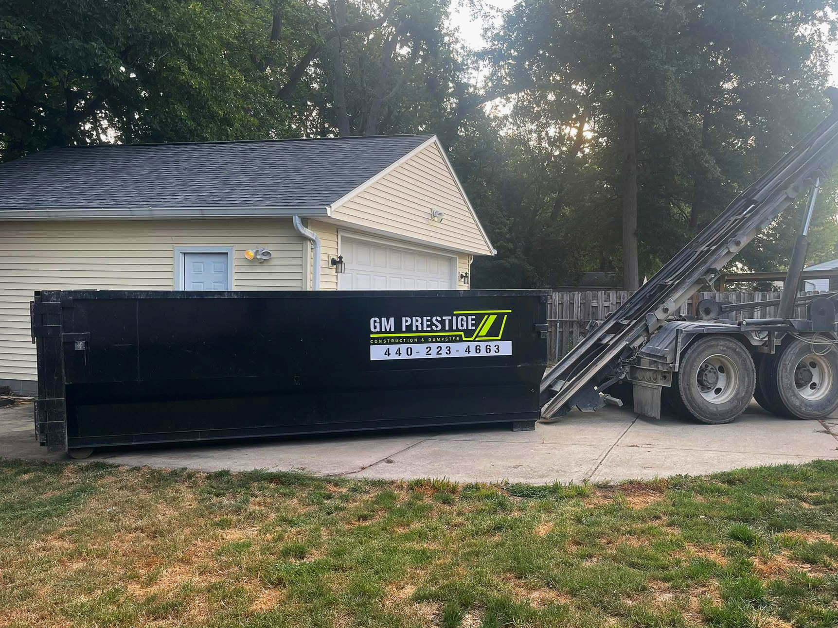 A black garbage container with a sign that says GM Prestige Construction & Dumpster along with a phone number, placed on a concrete driveway in front of a yellow garage with a door and a small door to a shed, in a backyard with trees and a wooden fence.