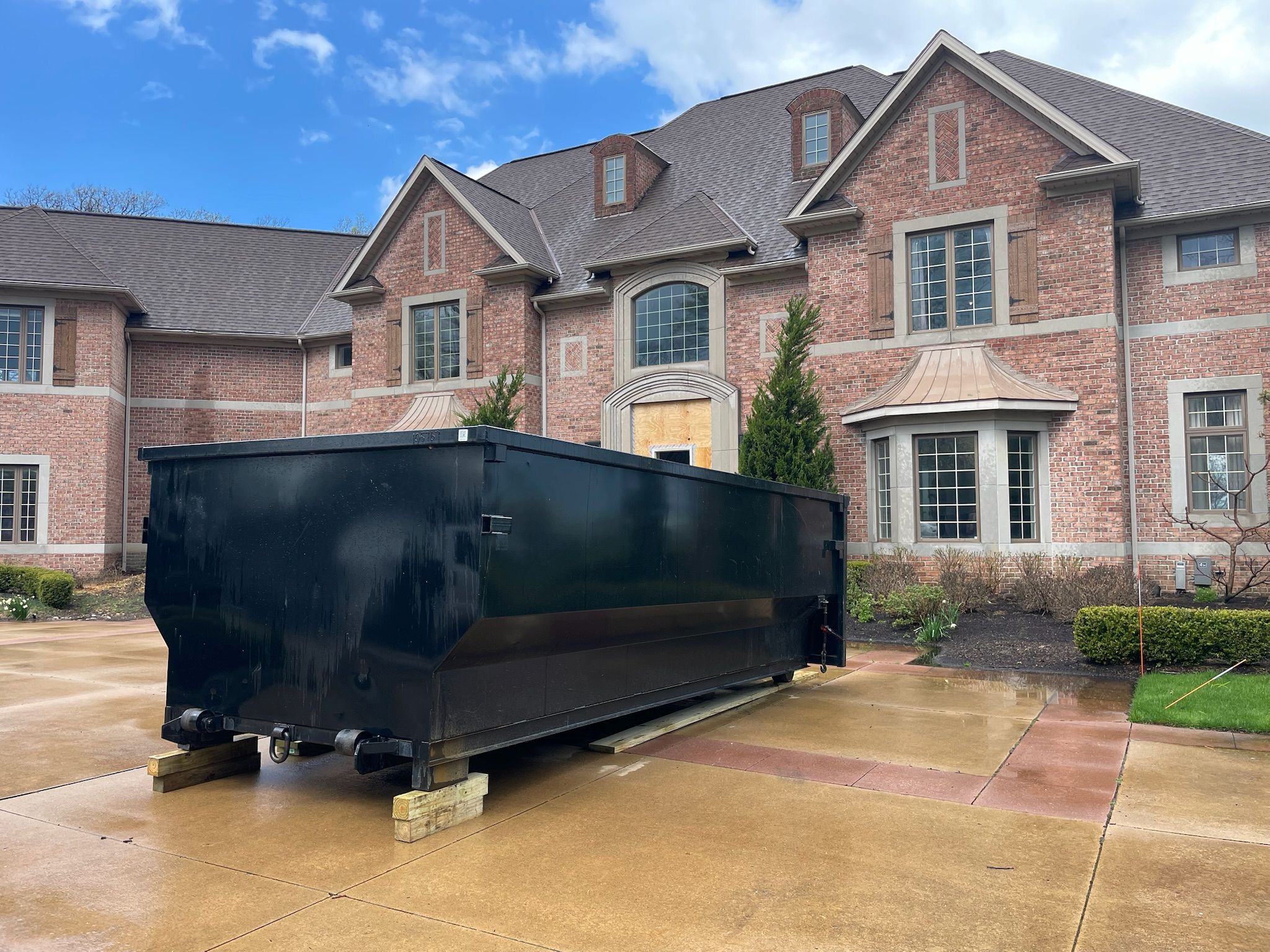 Black dumpster on a wet driveway in front of a large brick house with multiple windows and shrubs, under a partly cloudy sky.