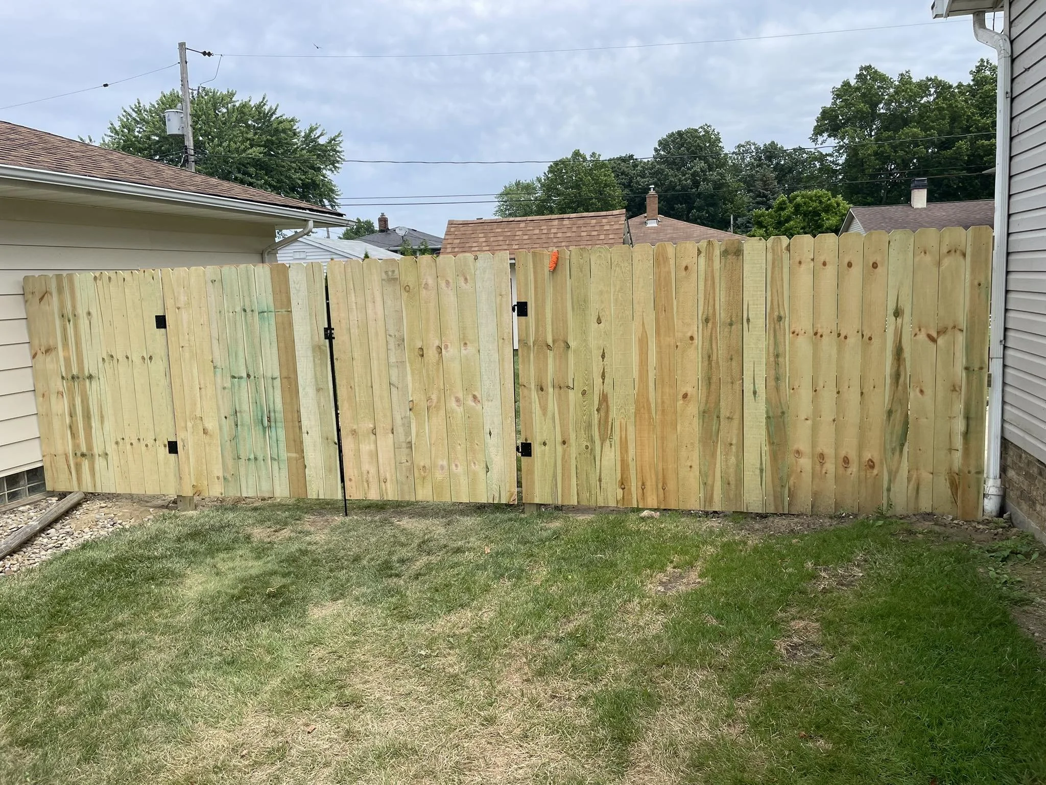 Wooden privacy fence in a backyard with grass, houses, and trees in the background under a cloudy sky.