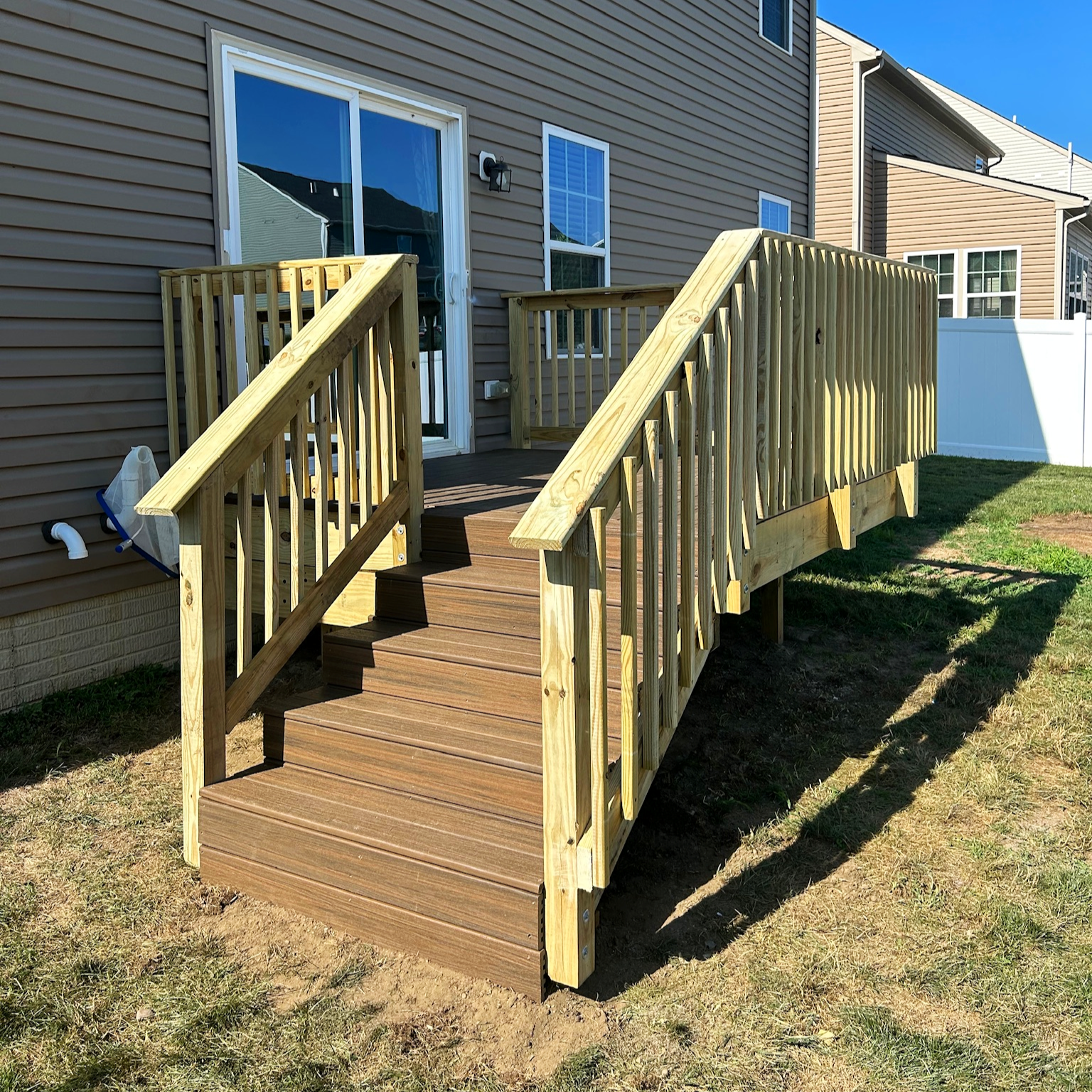 Newly built wooden deck with stairs and railings attached to beige house with sliding glass door, adjacent to a grassy yard and neighboring houses in bright sunlight.